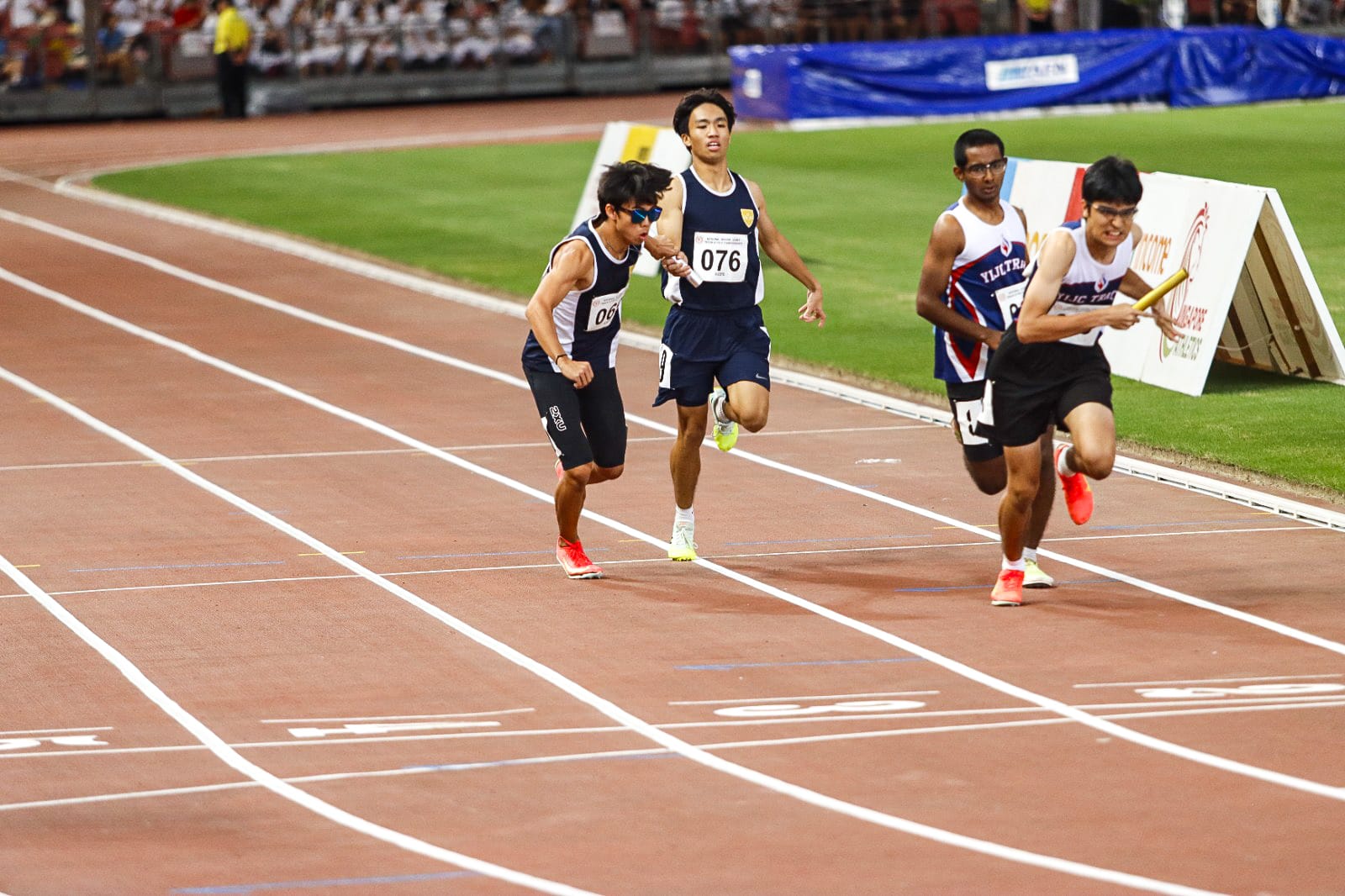 Four male runners compete in a relay race on a red track, one holding a baton mid-handoff.