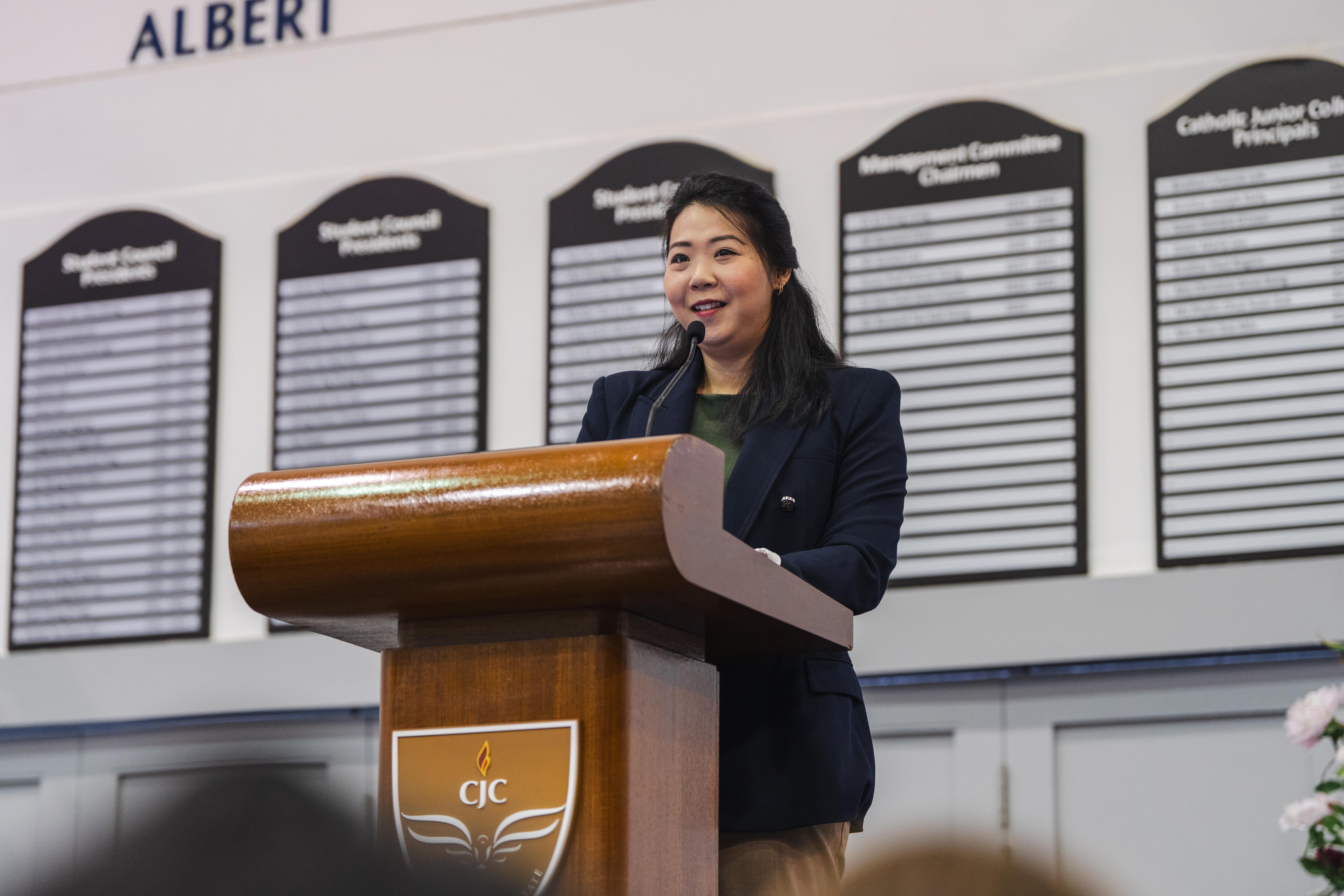 A smiling woman in a navy blazer speaks at a wooden CJC podium with honor boards on the wall behind her.