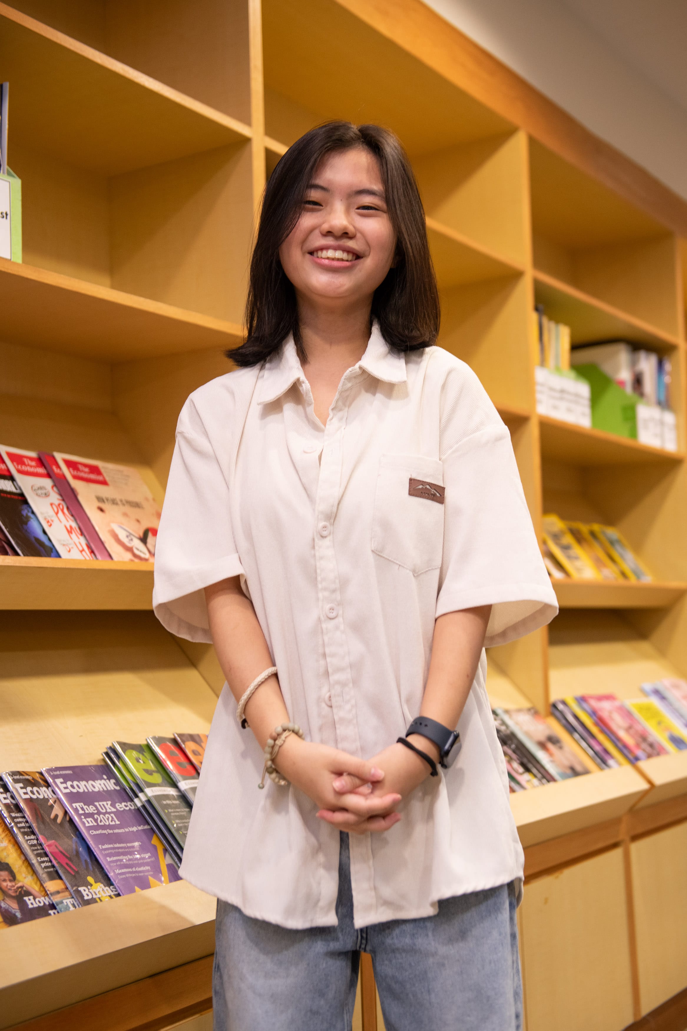 Young woman with dark hair smiling, wearing a white shirt and jeans, standing in front of wooden shelves with books.