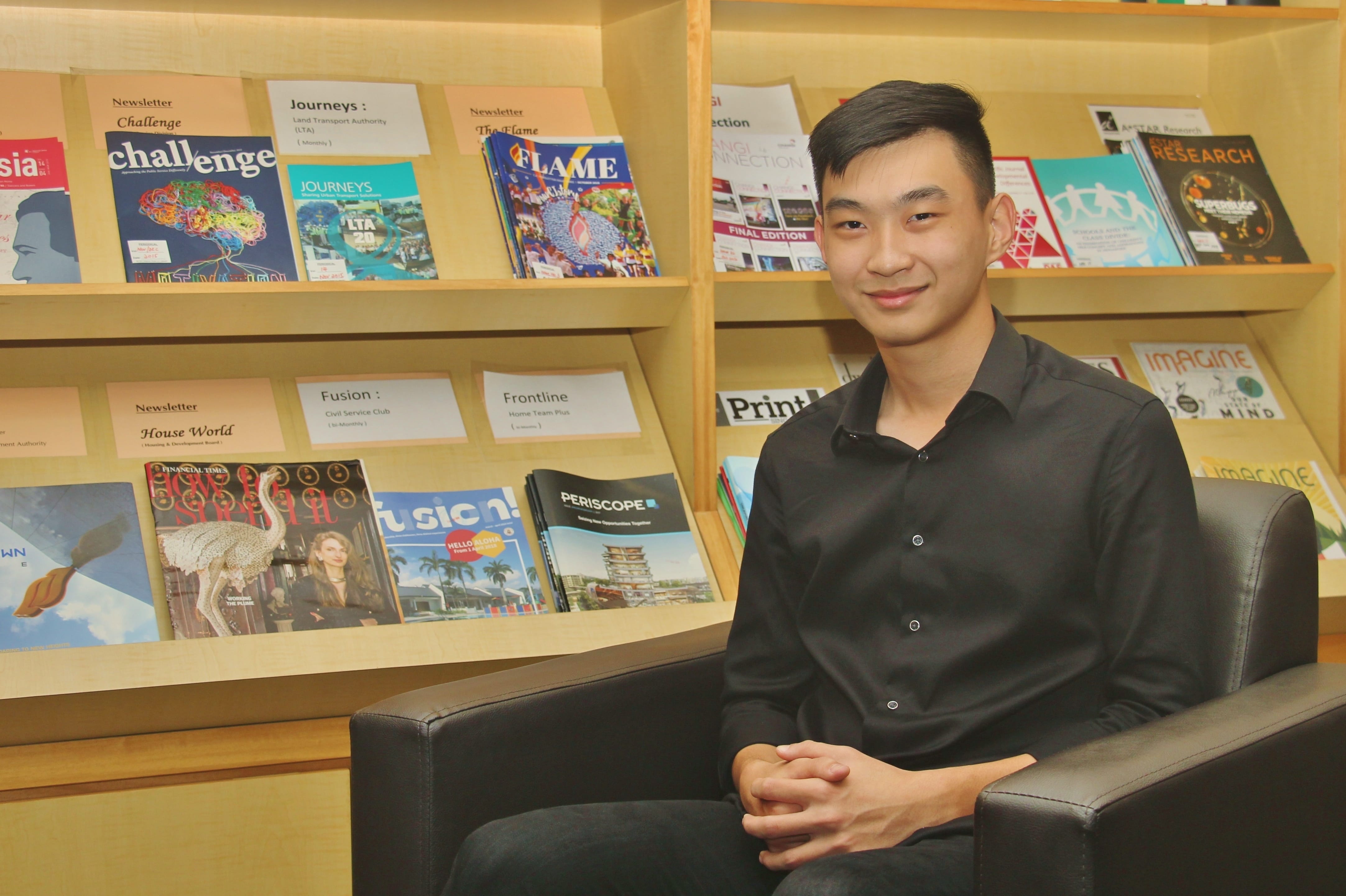 Man in a black shirt sits in an armchair in front of wooden shelves filled with magazines.