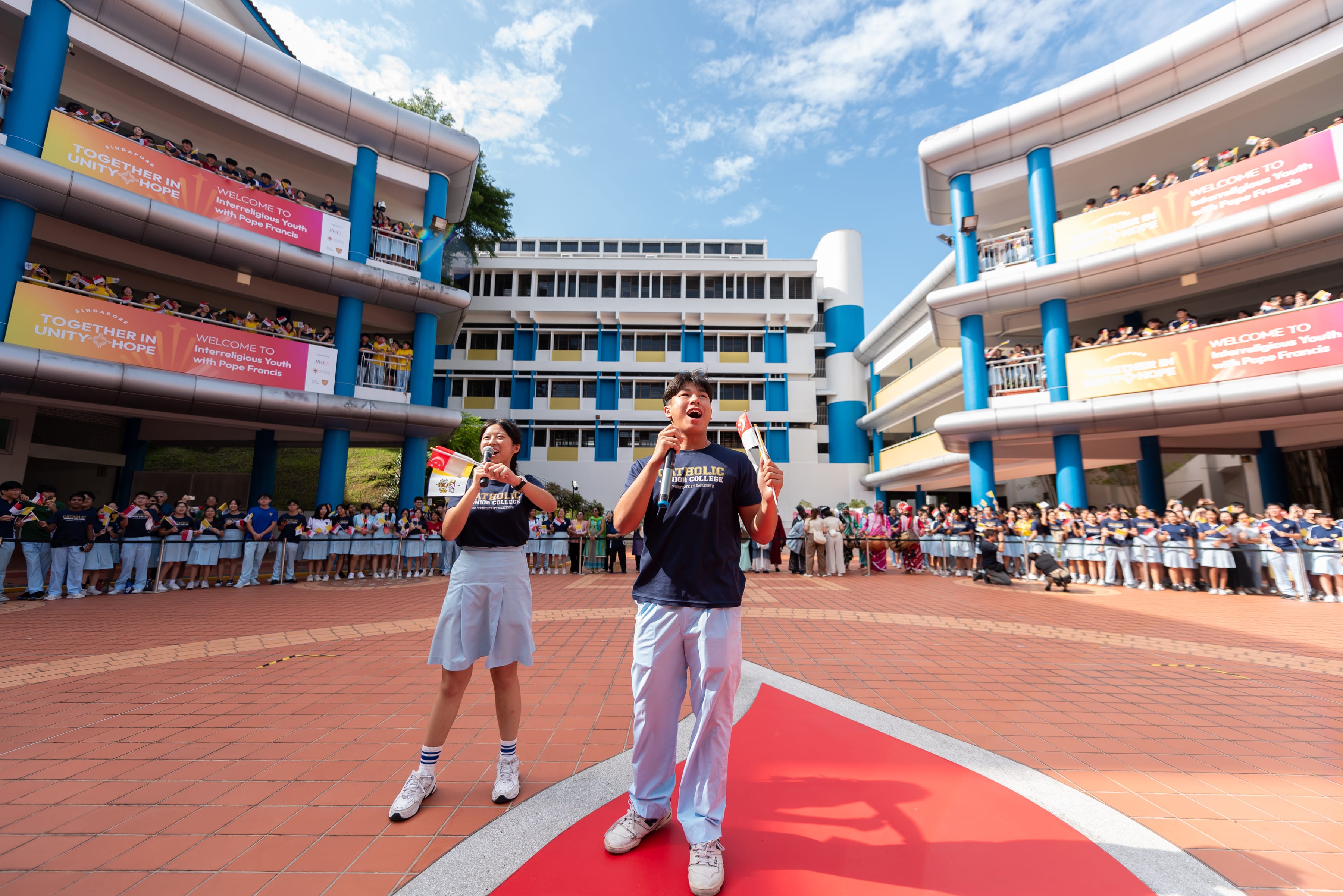 Two students speak in a school courtyard as crowds watch from balconies and ground level under blue sky.