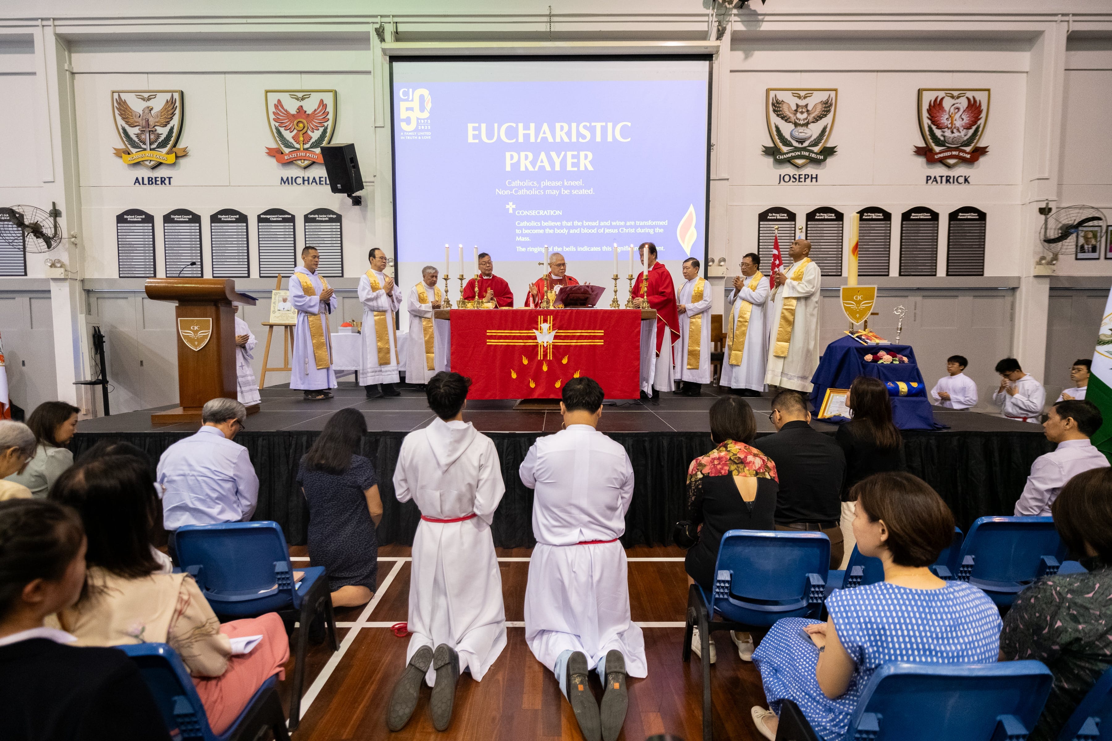 Catholic Mass with priests at a red altar on stage, congregation kneeling, and a screen showing Eucharistic Prayer.