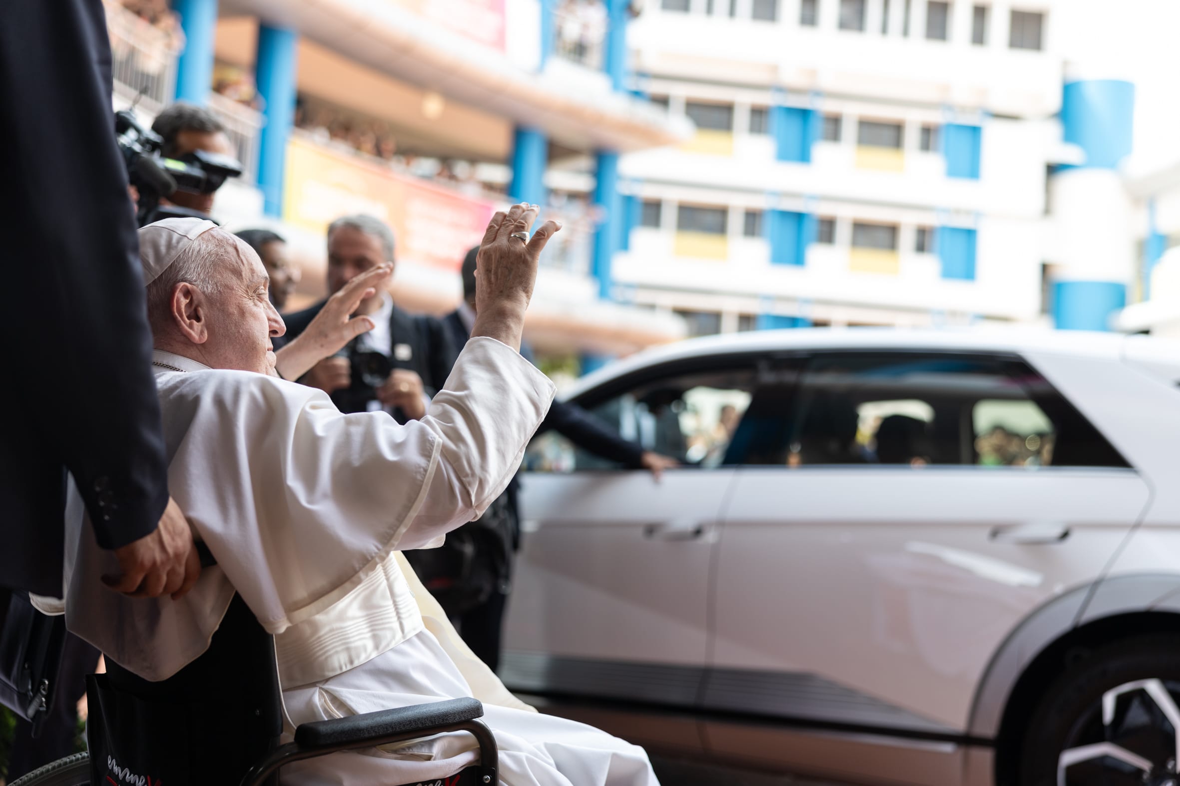 Elderly man in white papal robes sits in a wheelchair, raising both hands in a greeting gesture near a white car.