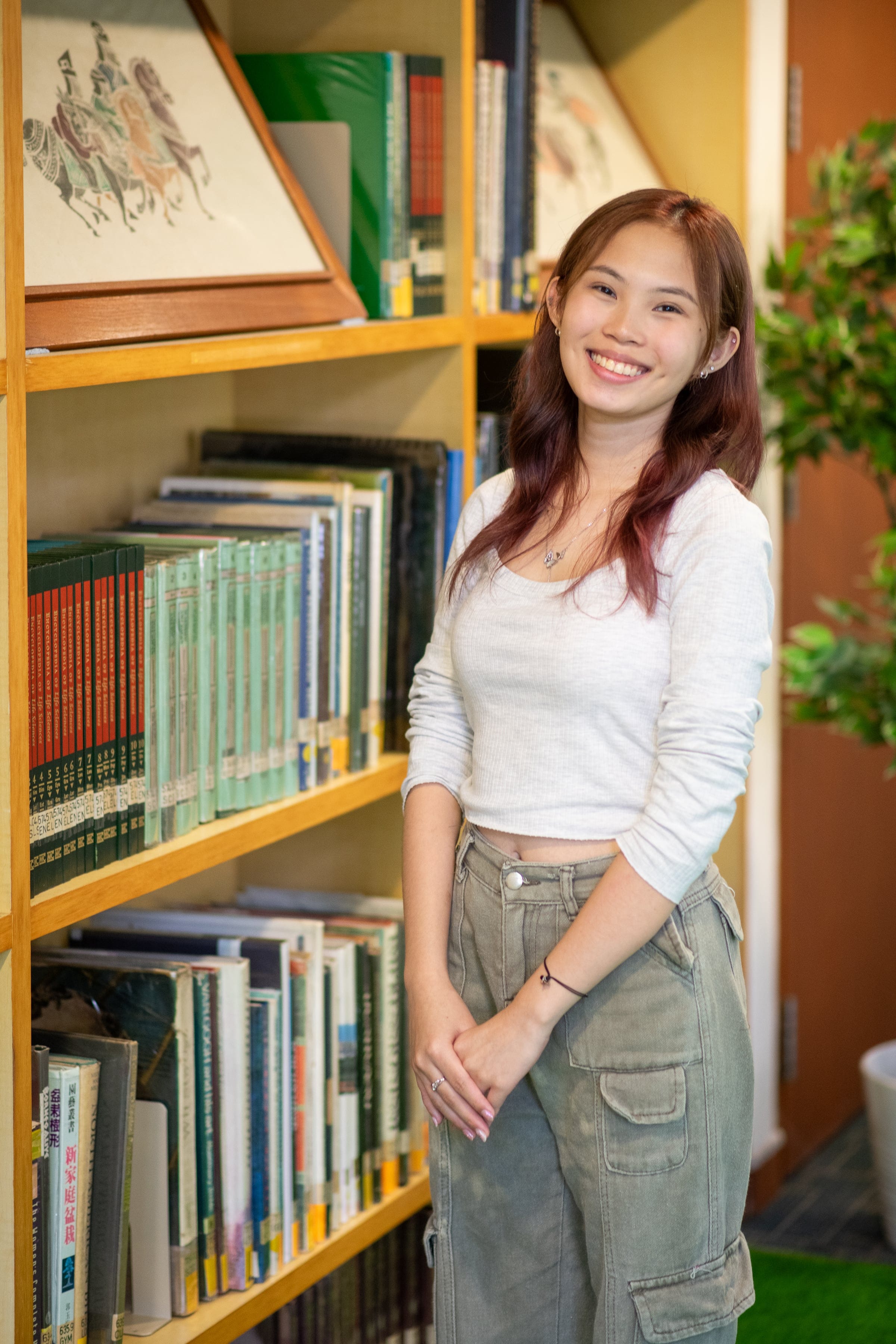 Young woman with long reddish hair, smiling, wearing a white crop top and gray cargo pants, standing beside a wooden bookshelf.