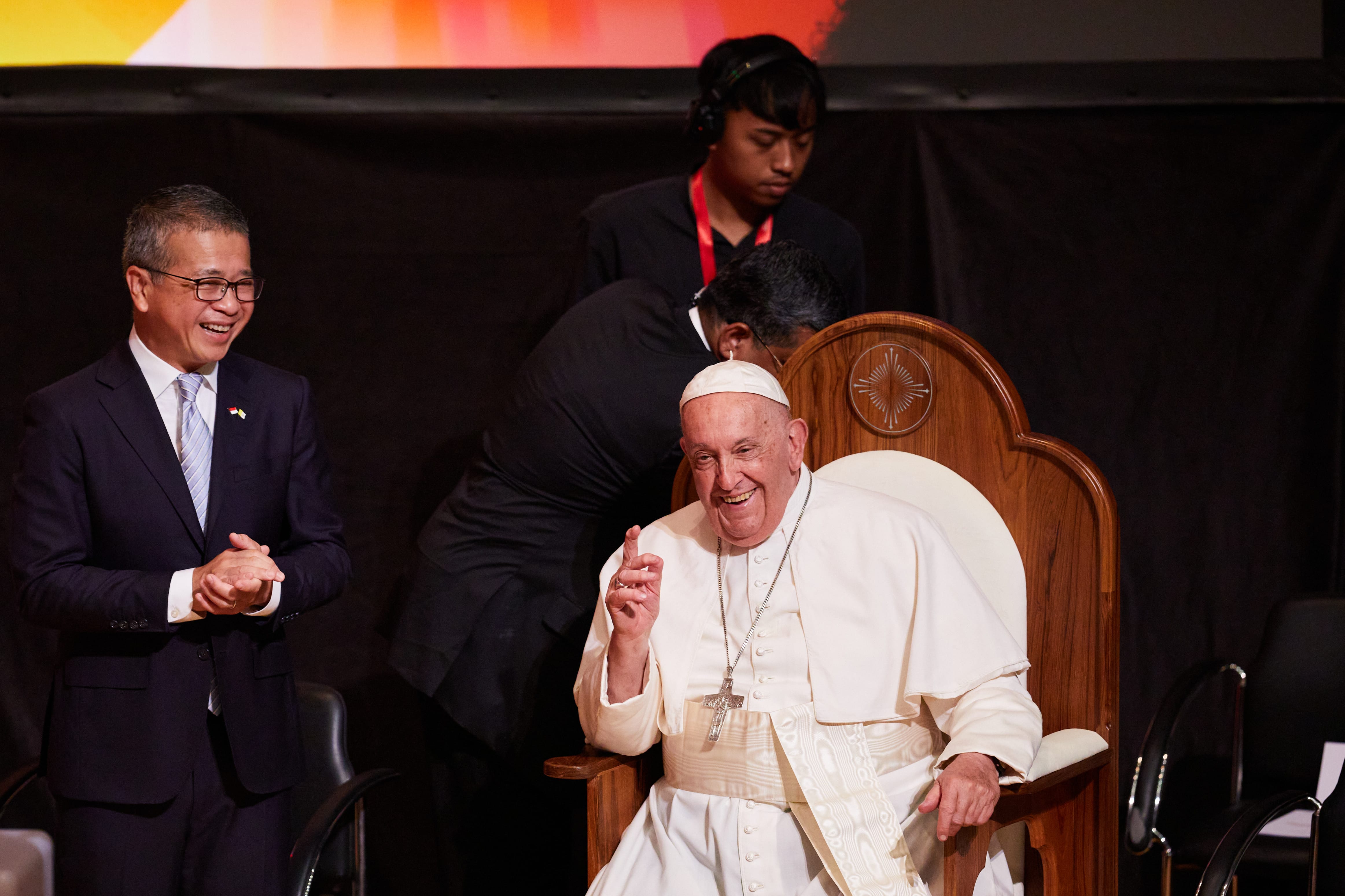 An elderly man in white papal robes and skullcap sits on a wooden throne, smiling and raising one finger, beside a laughing man in a dark suit.