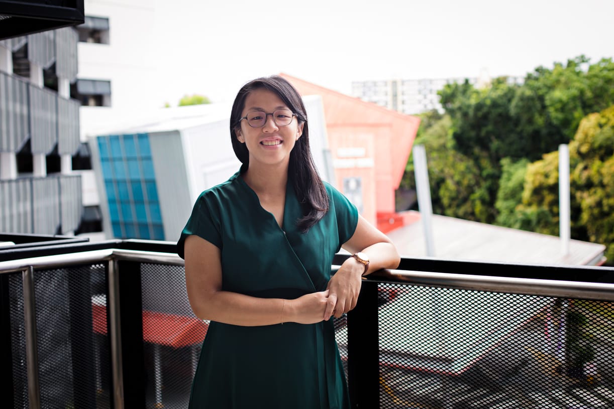 A smiling woman with glasses in a green dress leans on a metal balcony railing with buildings and trees behind her.