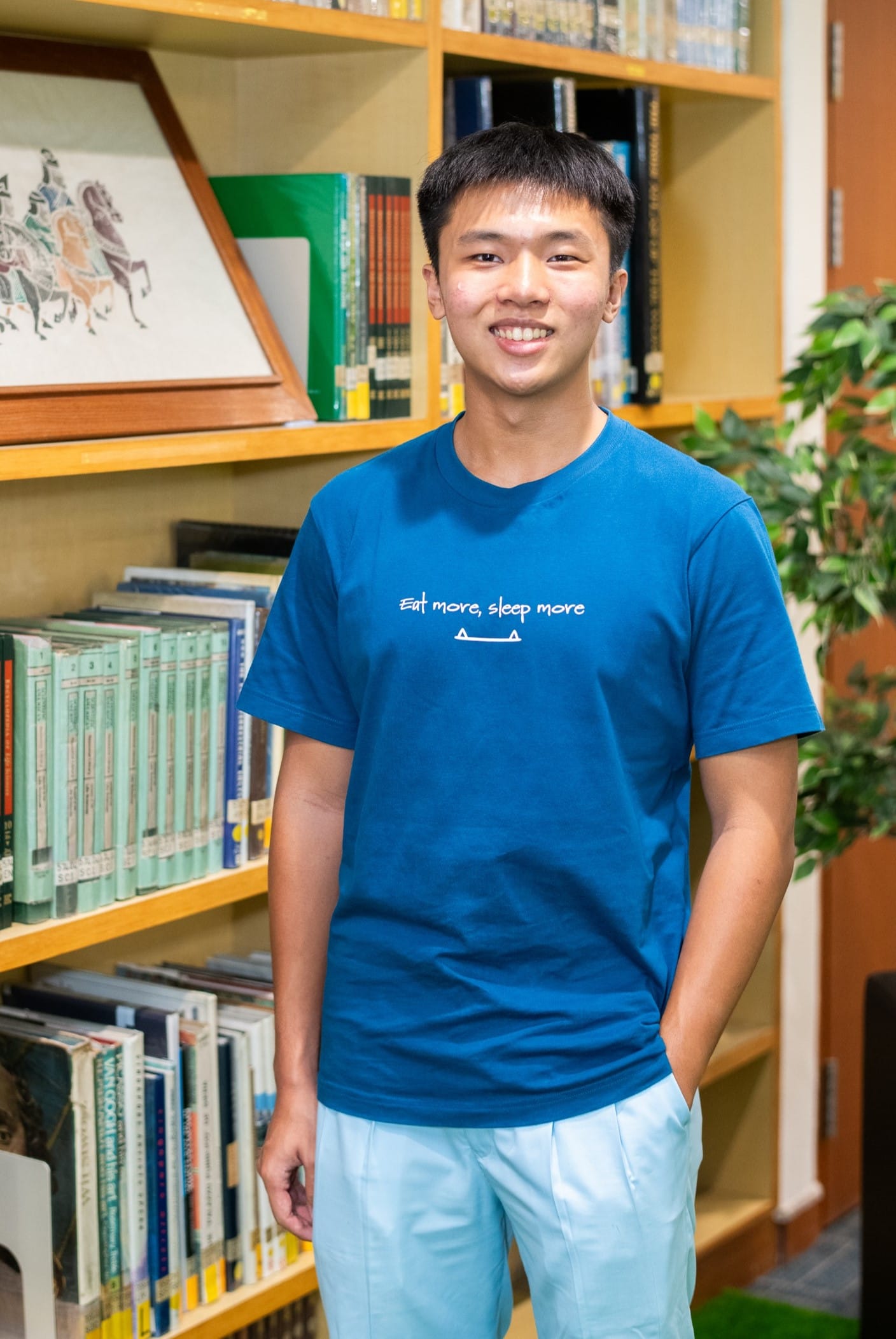 Young man smiling, wearing a blue "Eat more, sleep more" t-shirt, standing in front of wooden bookshelves.