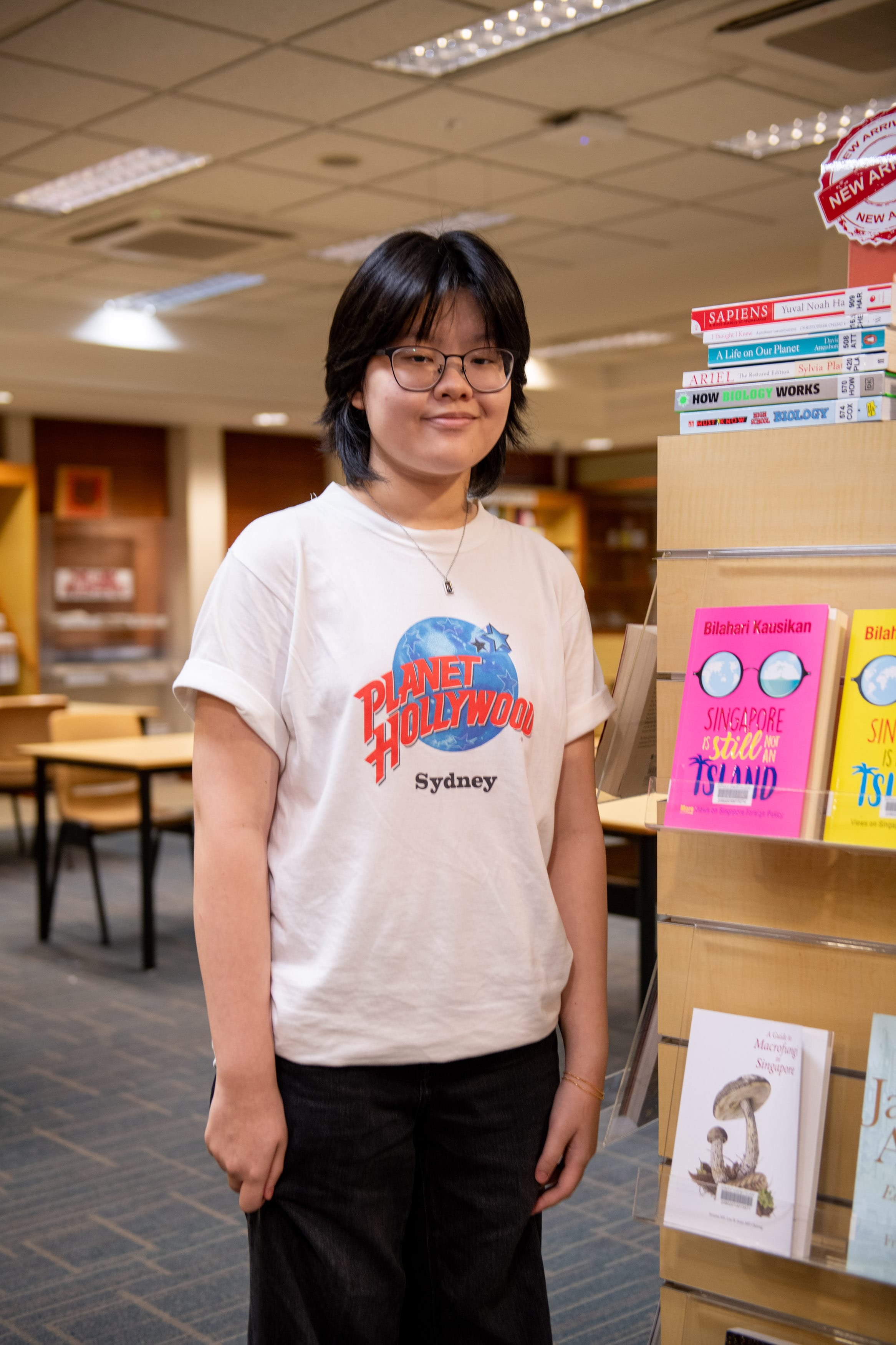 Young person with glasses wearing a Planet Hollywood Sydney t-shirt standing beside a bookshelf in a library.
