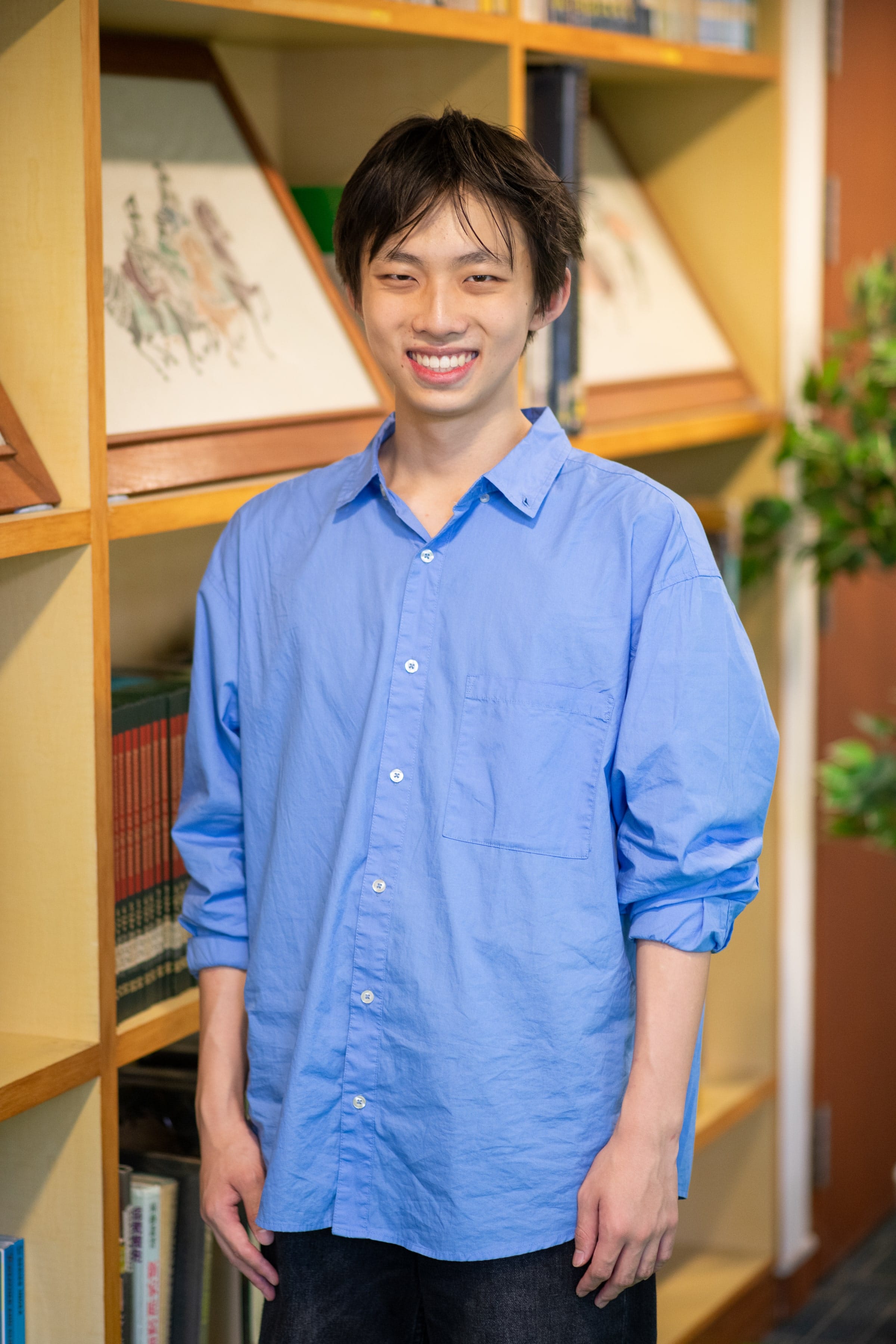 Young man with short dark hair smiling, wearing a blue button-up shirt, standing in front of a wooden bookshelf.