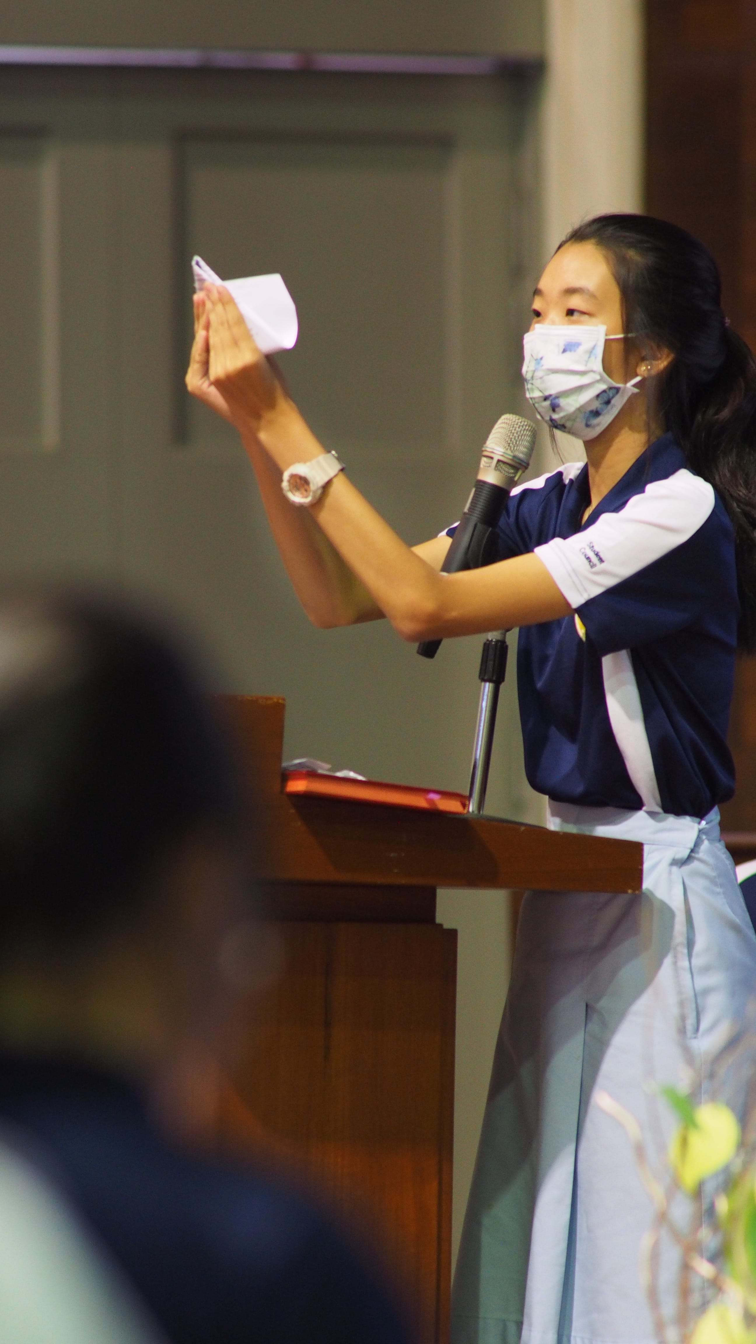 A young woman wearing a face mask speaks at a podium microphone, holding up a small piece of paper.