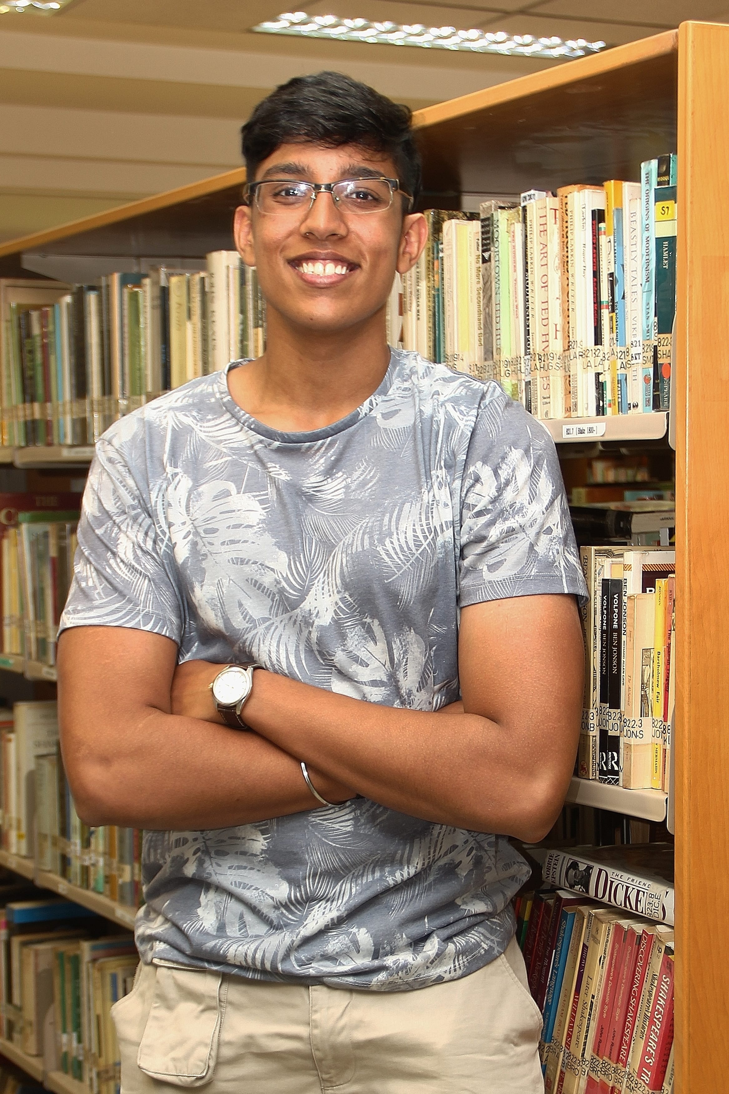 Smiling person with glasses and crossed arms stands by library shelves, wearing a gray patterned shirt.