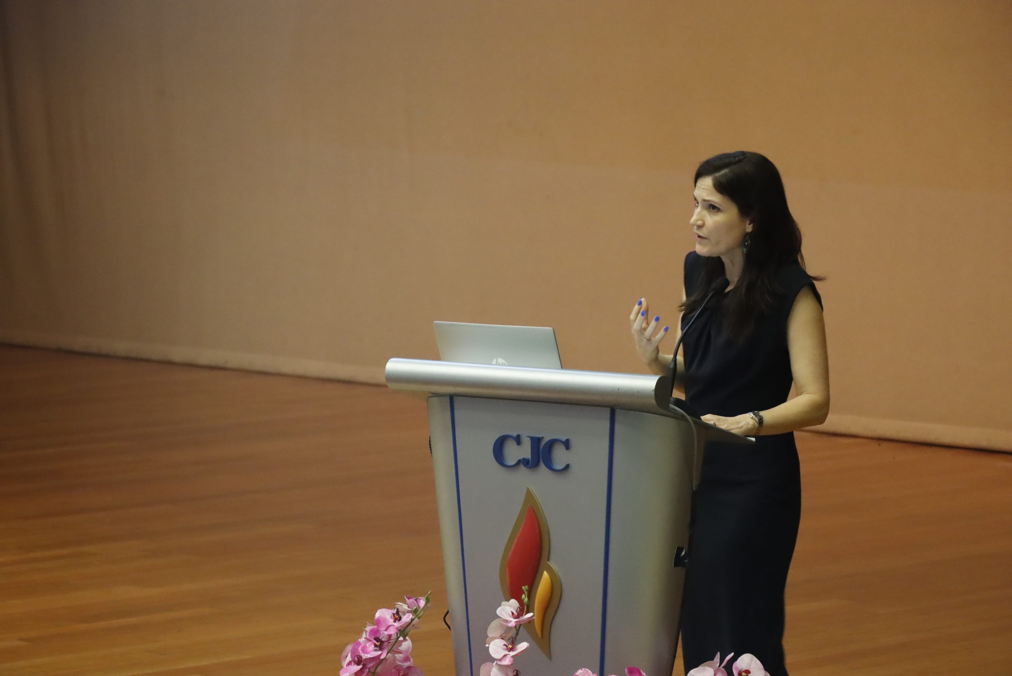 Woman in black dress speaking at a CJC podium with a laptop, pink flowers at the base, on a wooden stage.