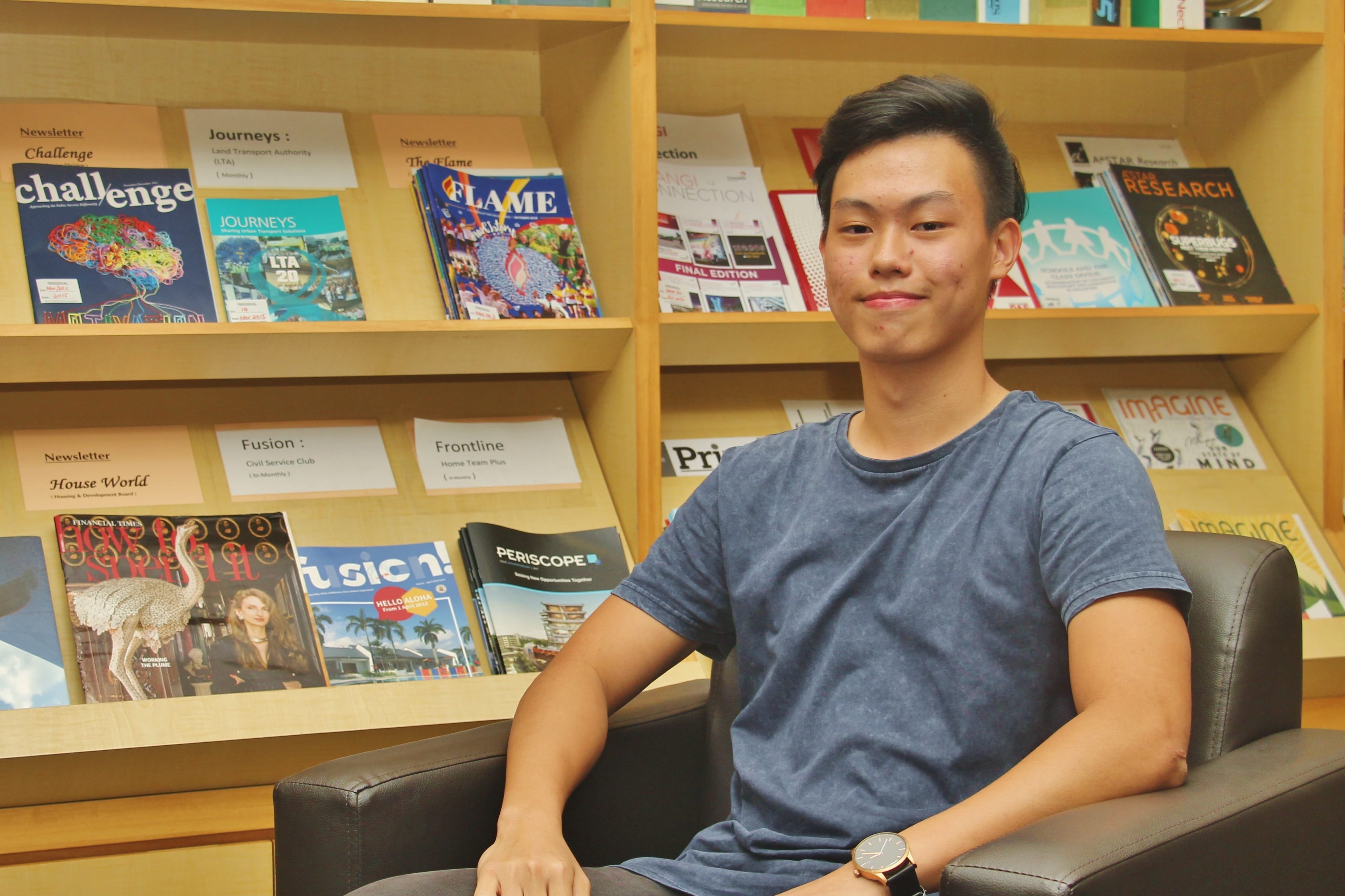 Young man in a blue t-shirt sitting in a chair in front of wooden shelves displaying various magazines.