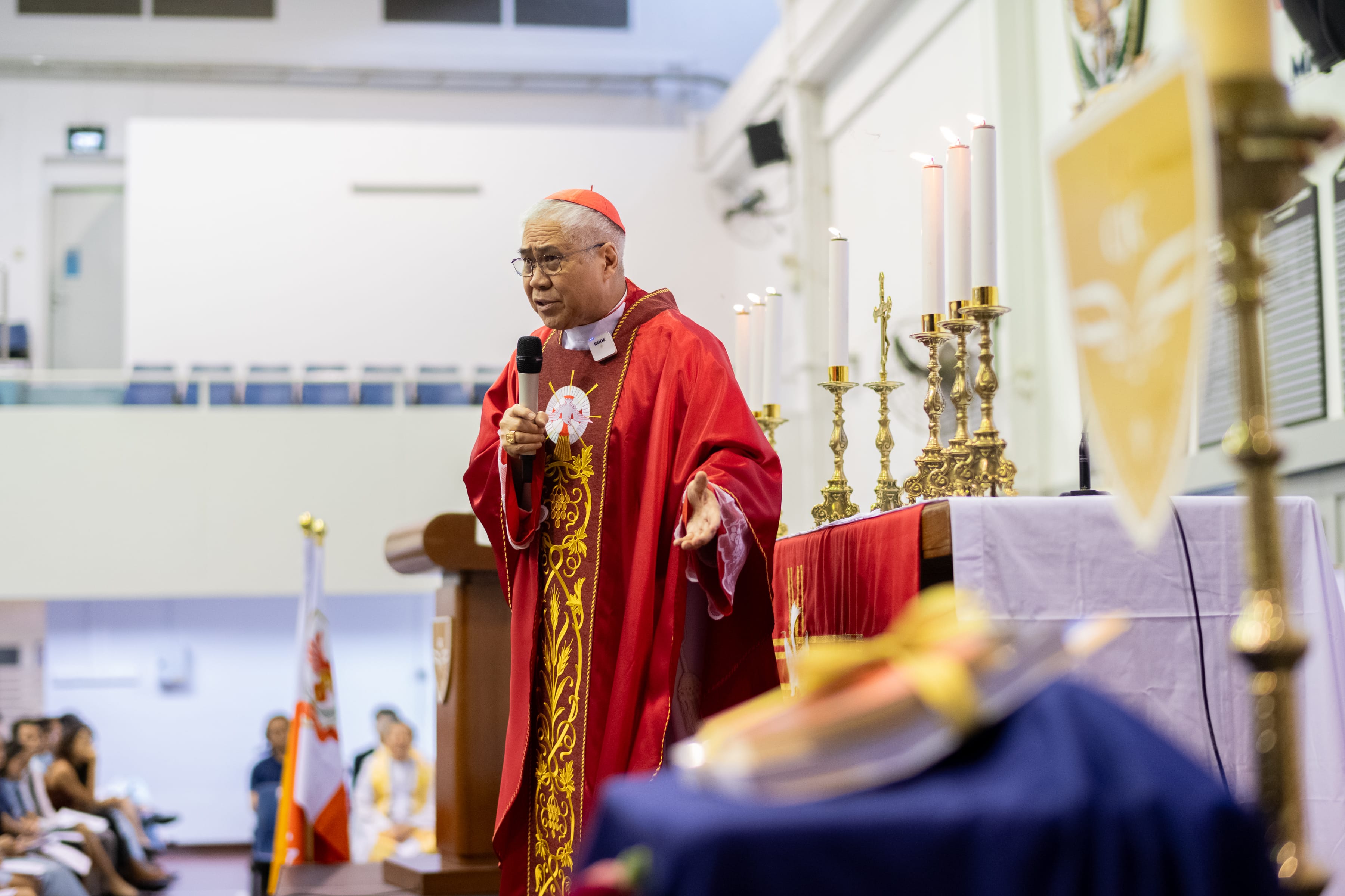 Clergyman in red robes and skullcap speaks into a microphone beside an altar with lit candles.