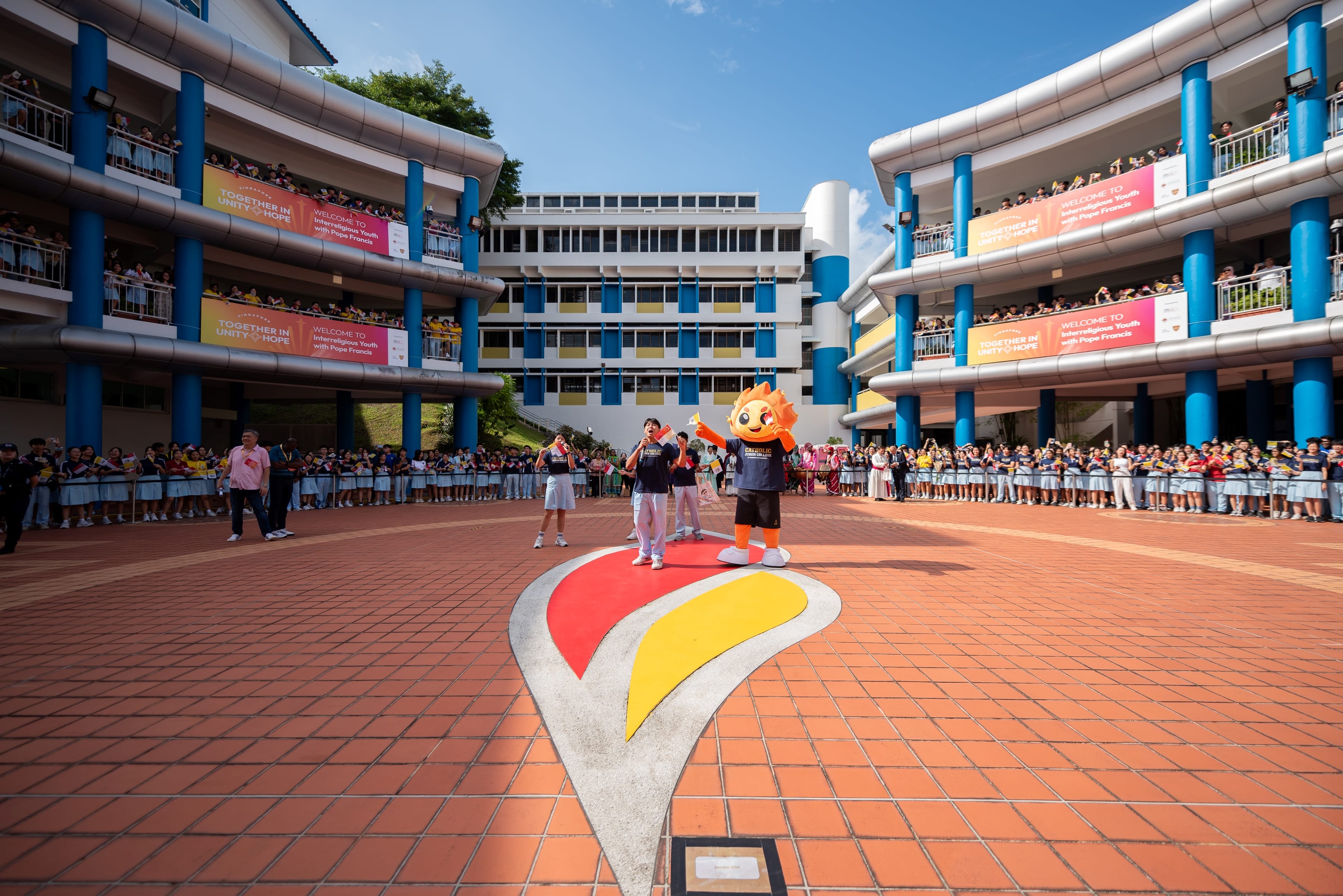 School courtyard with crowds on balconies; mascot and students stand on a flame-shaped emblem on bricks.