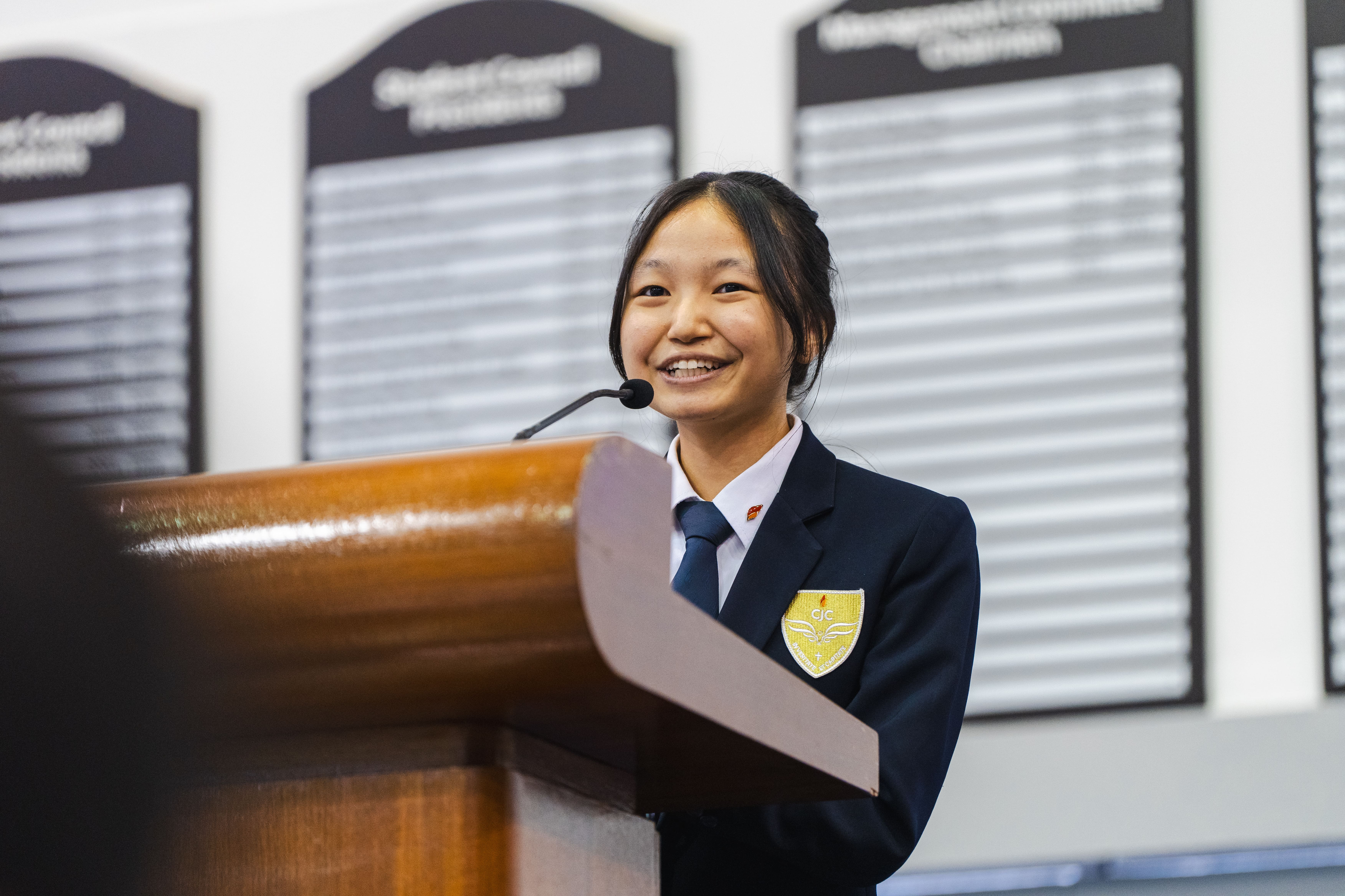 A smiling young girl in a school blazer with a badge speaks at a wooden podium with a microphone.