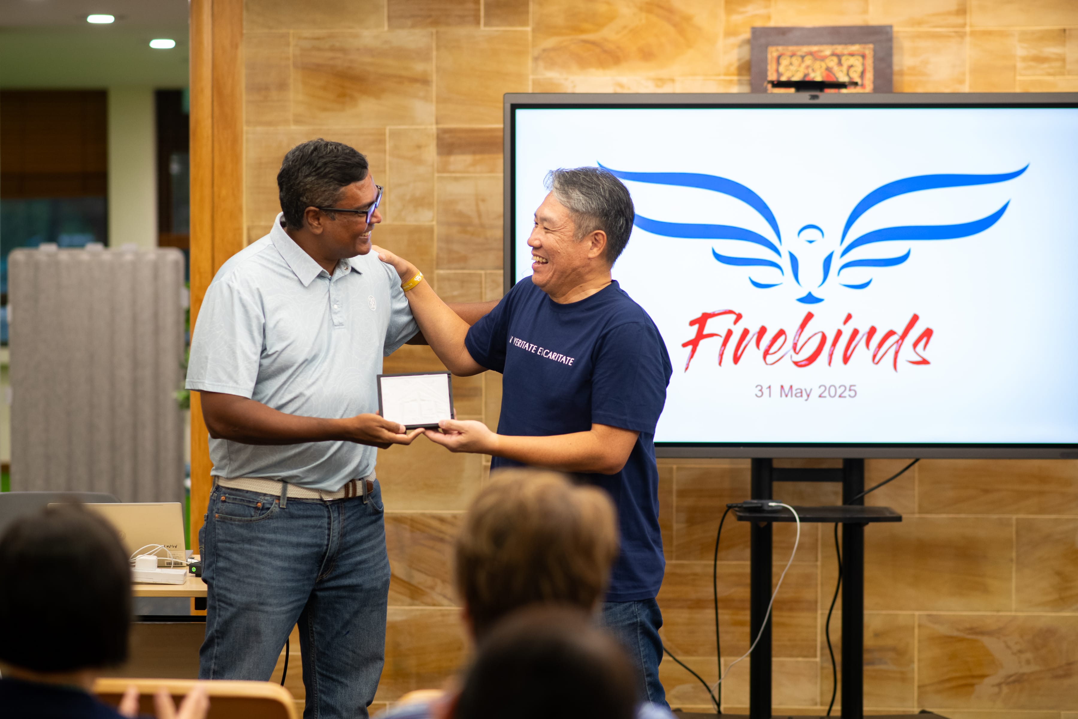 Two men smile as one presents a plaque; screen behind shows blue wings and Firebirds, 31 May 2025
