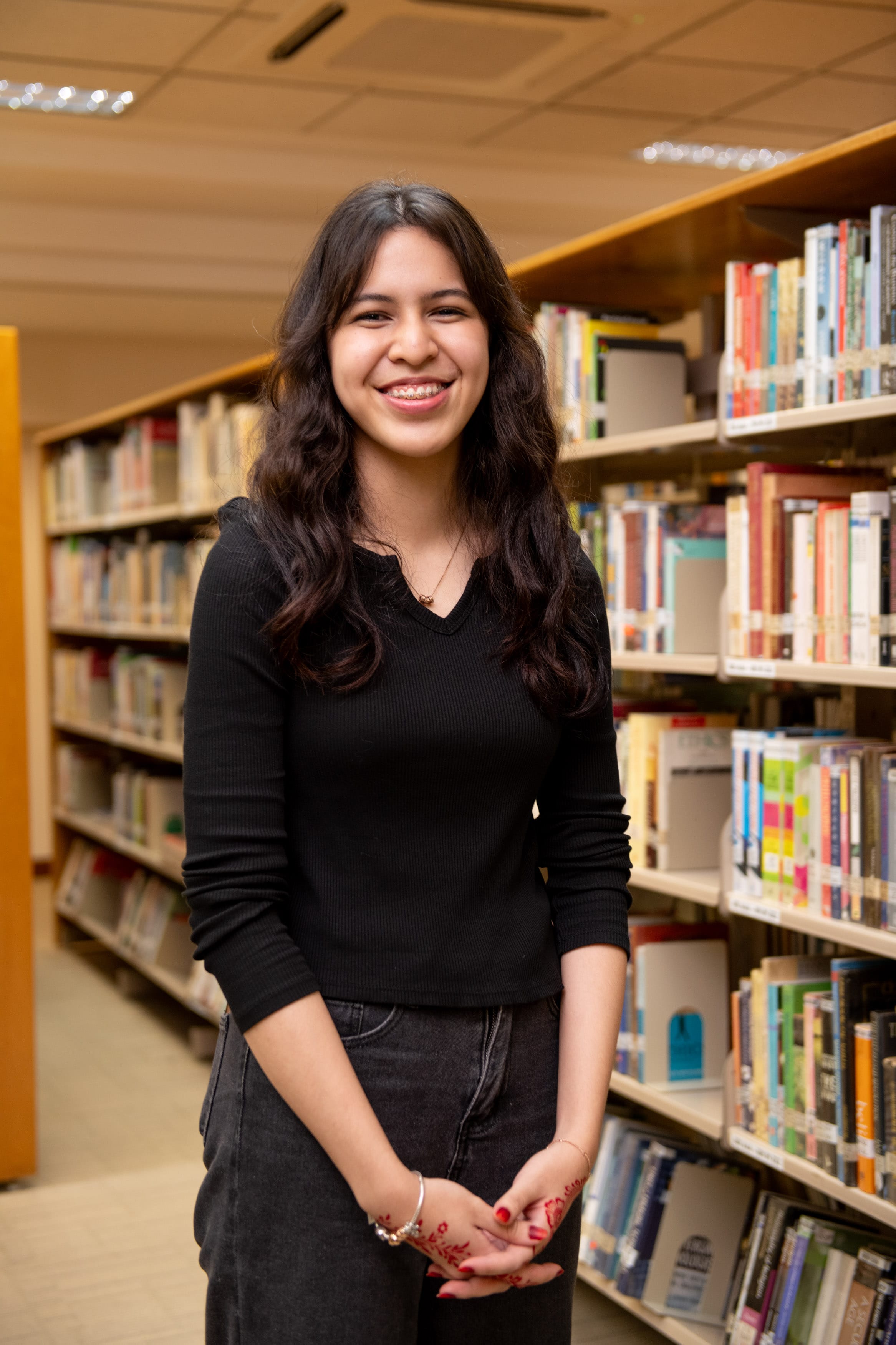 Young woman with long dark hair smiling in a library aisle, wearing a black top and dark jeans.