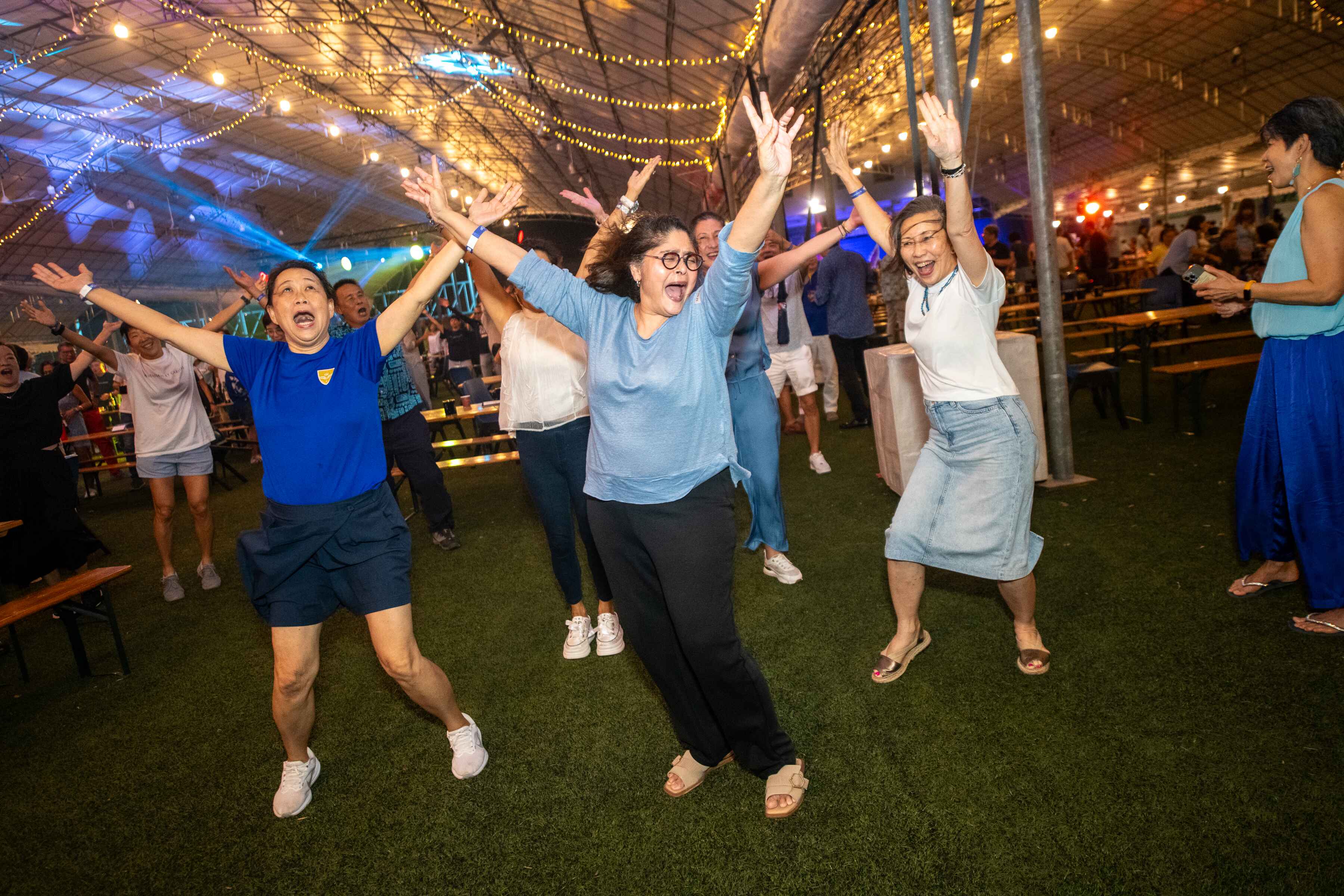 A group of joyful people dancing with arms raised at an indoor event under string lights on a green turf floor.