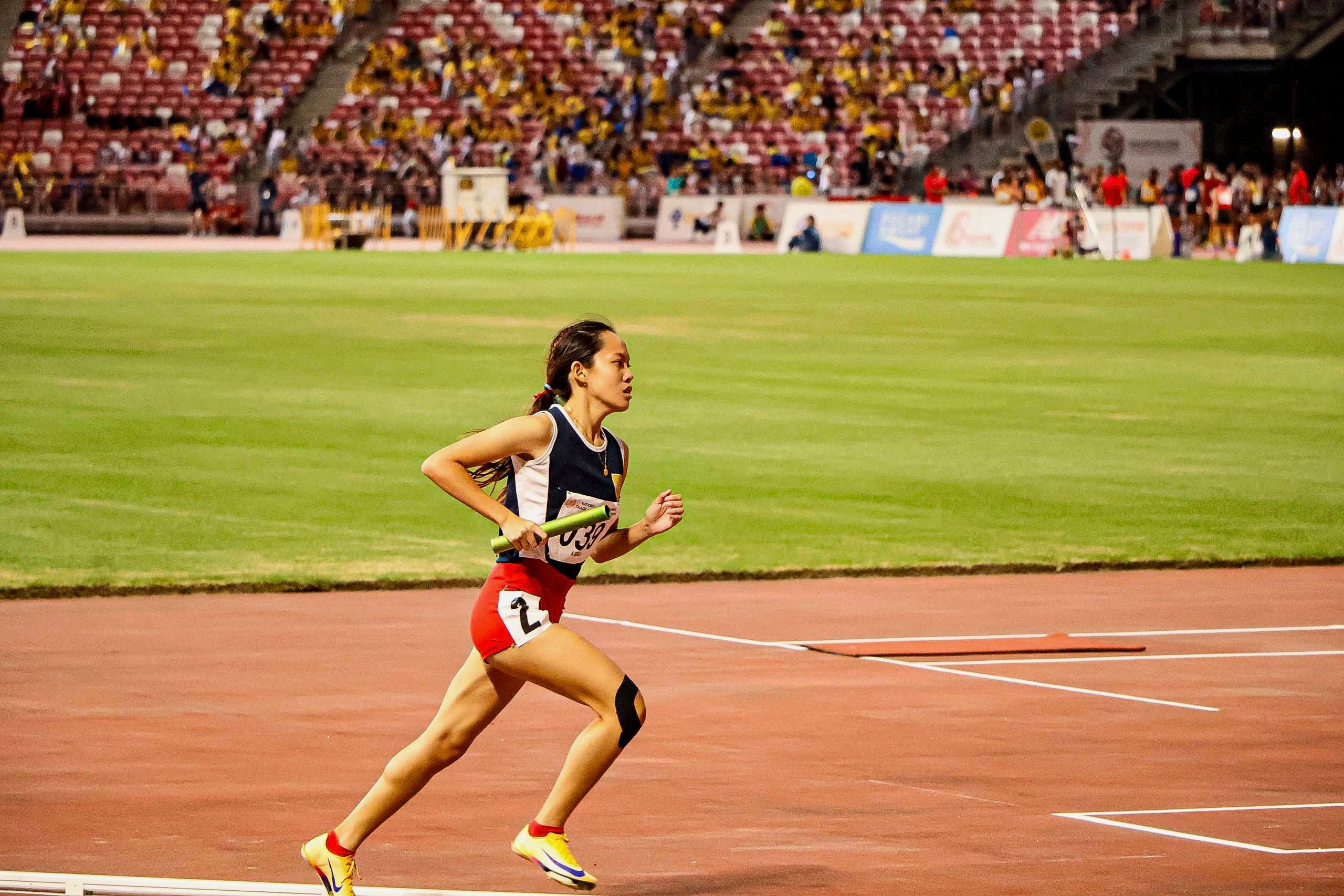Female relay runner holding a green baton sprints on a red track in a stadium with spectators in the stands.