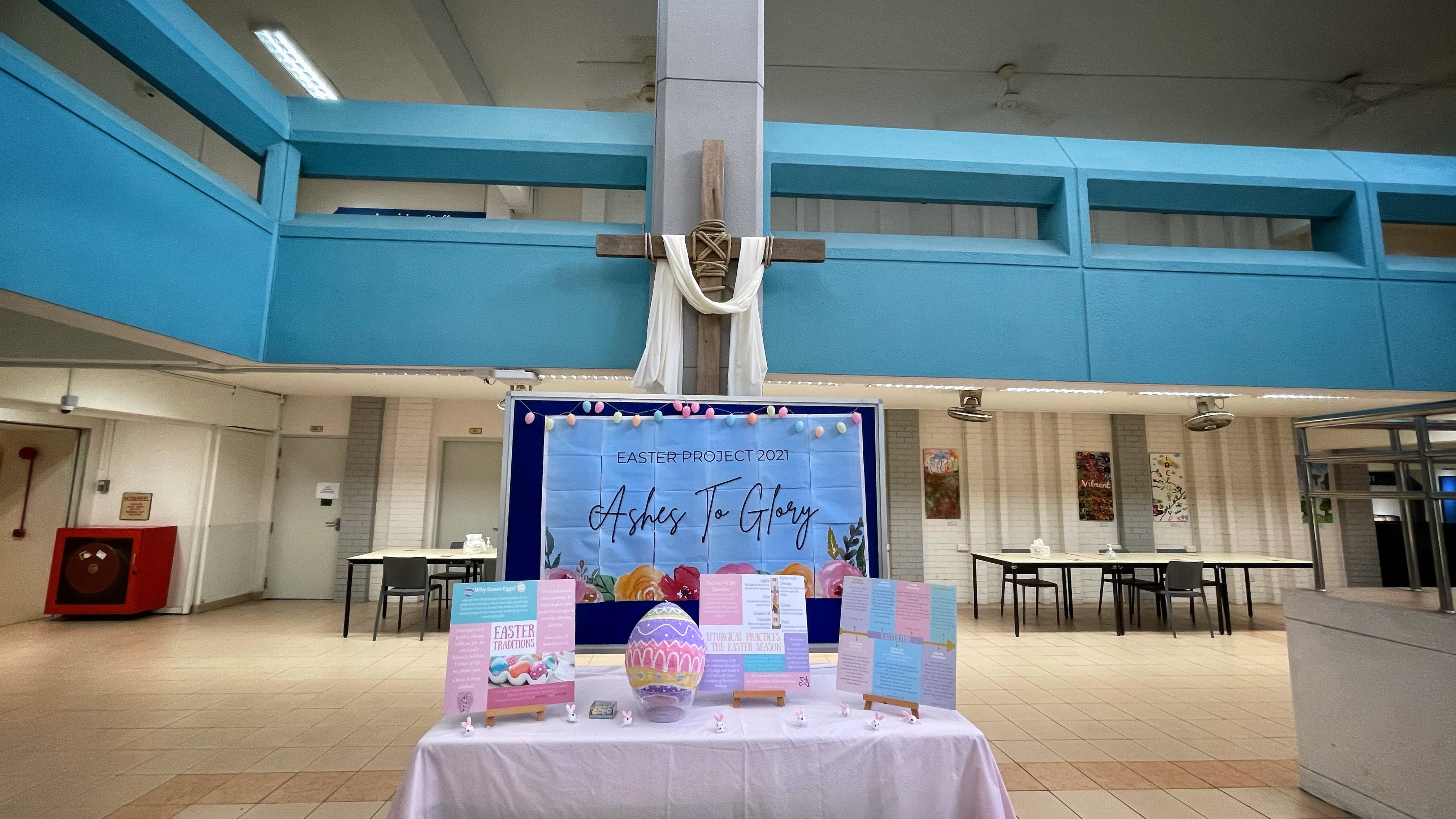 A school hall Easter 2021 display with an "Ashes To Glory" banner, wooden cross, decorated egg, and info boards on a table.