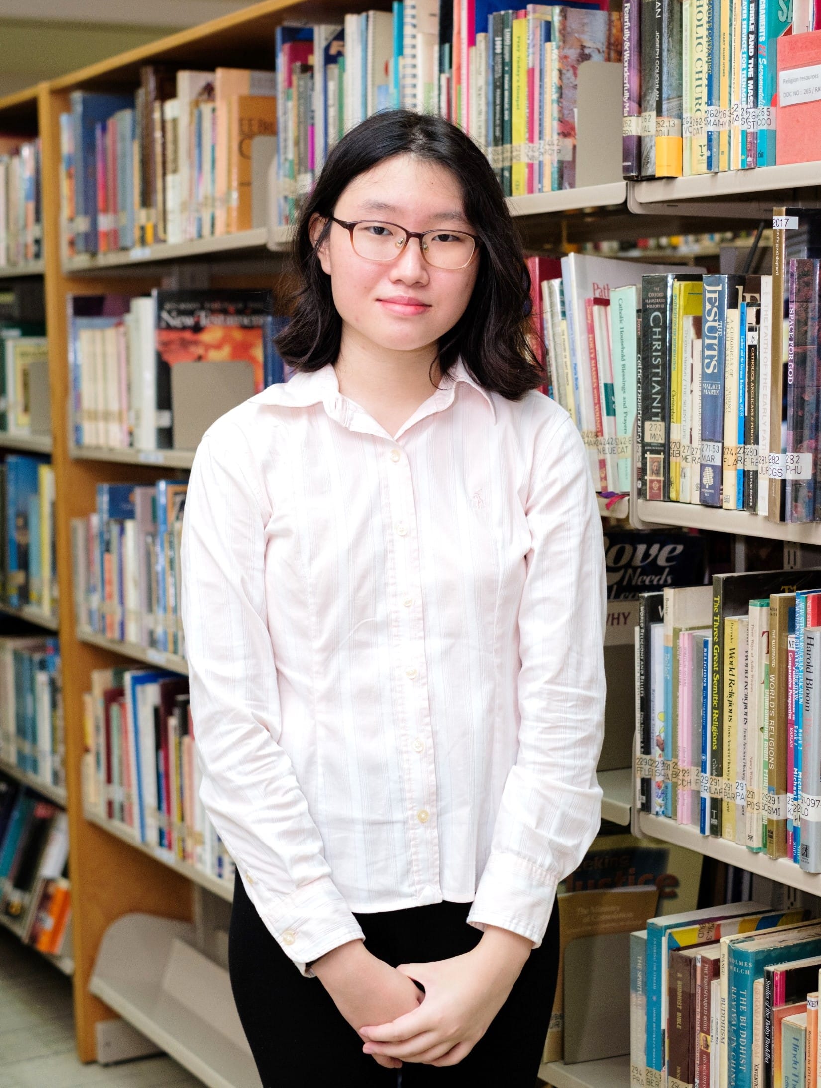 Young woman with glasses and dark hair wearing a pink shirt stands between library bookshelves, smiling.