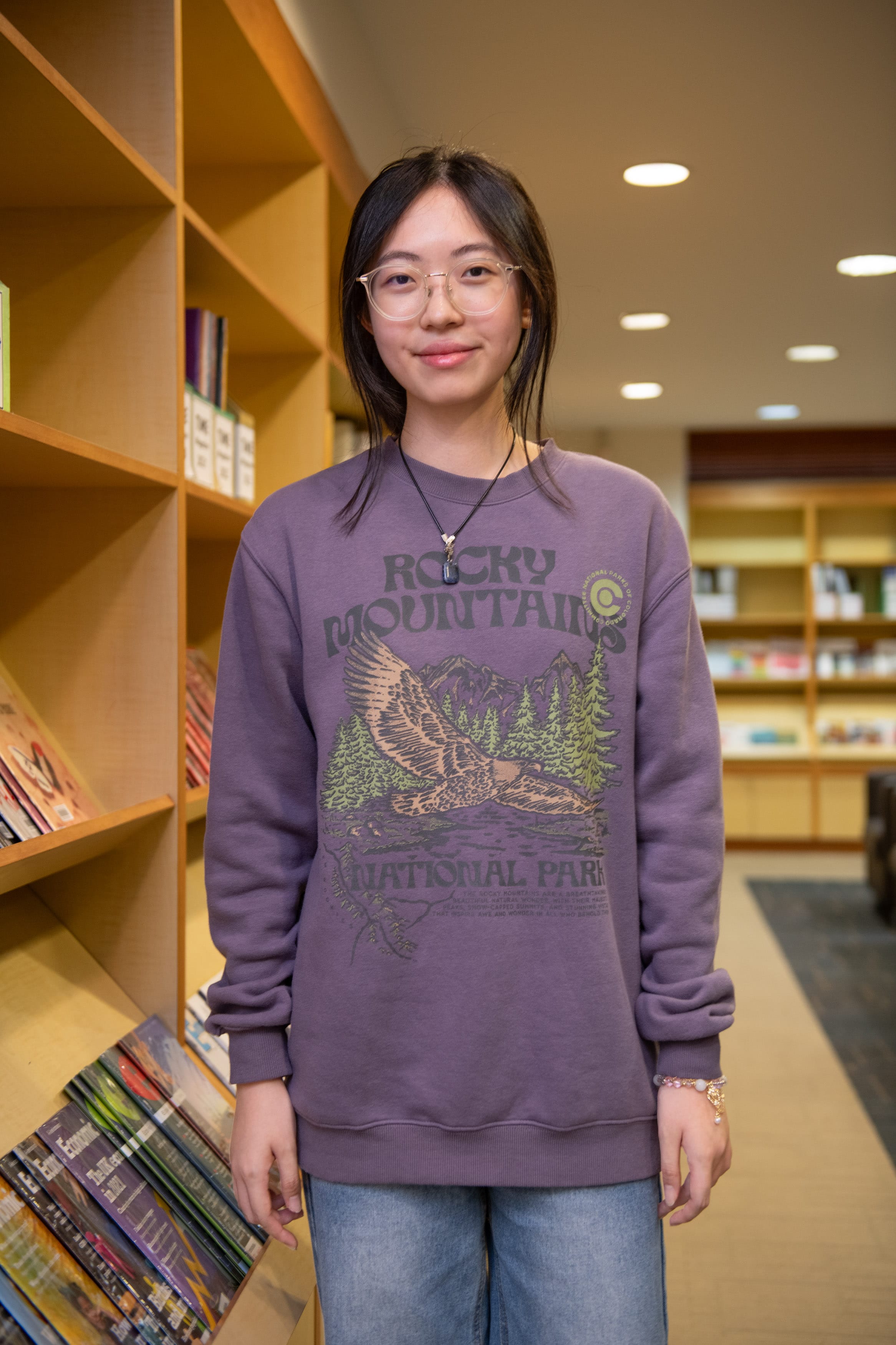 Young woman with glasses wearing a purple Rocky Mountains National Park sweatshirt stands smiling in a library.