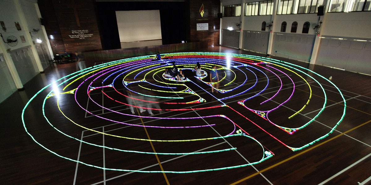 A large glowing neon labyrinth made of colorful light strips on a dark gymnasium floor with people walking inside.