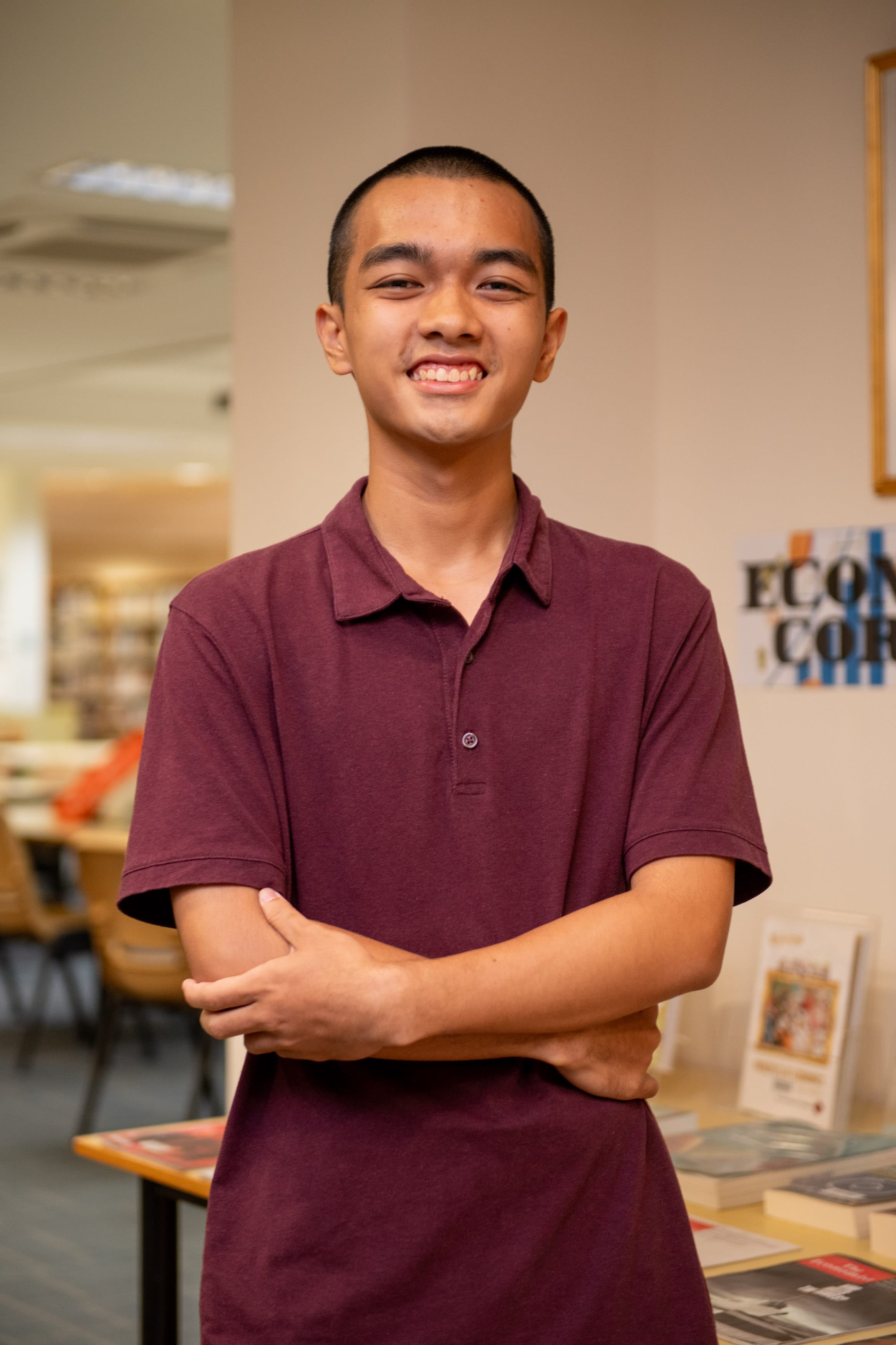 Young man in a dark red polo shirt smiling with arms crossed, standing in a library.