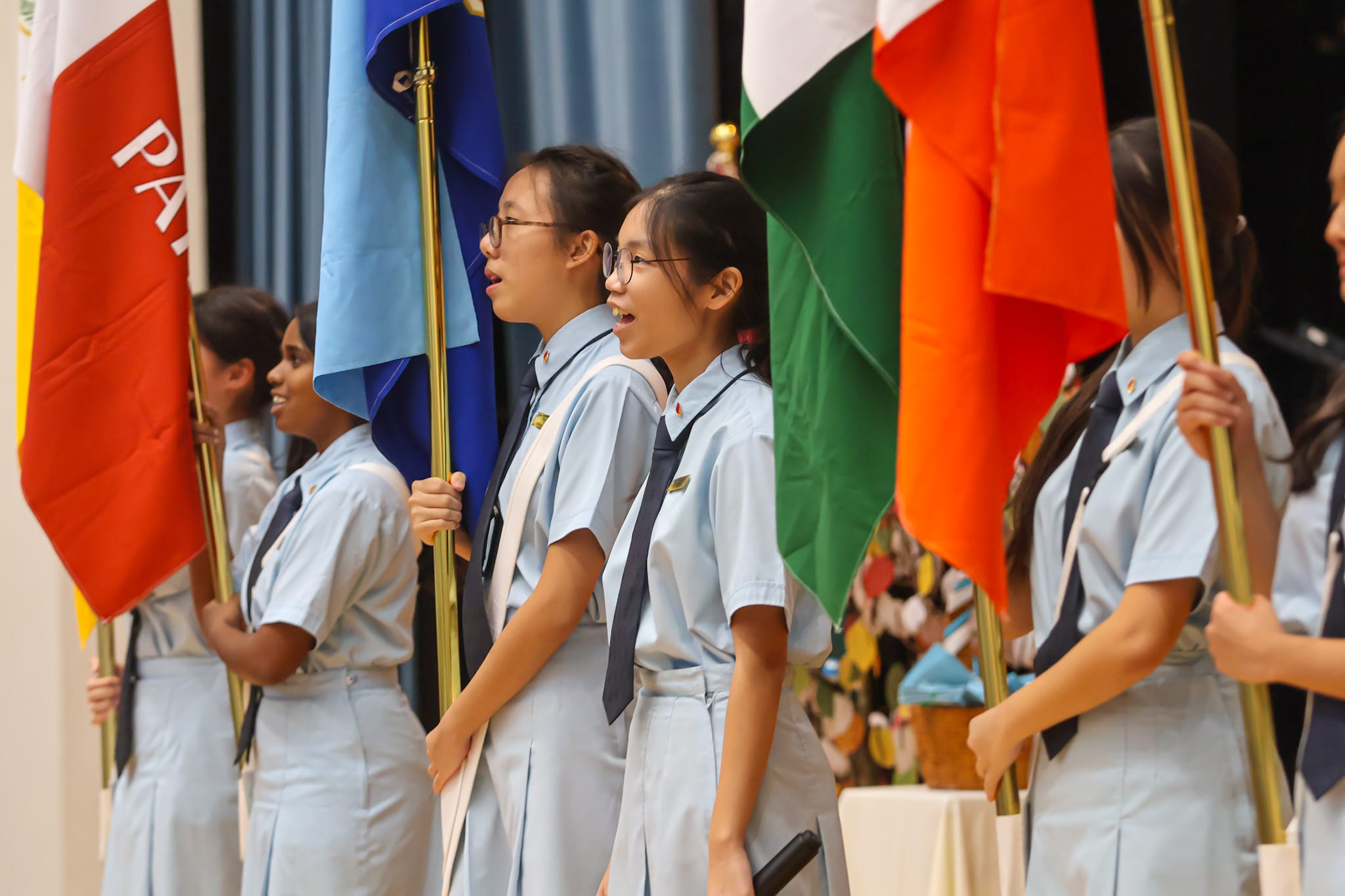 Students in light blue uniforms hold colorful international flags during a school ceremony on a stage.