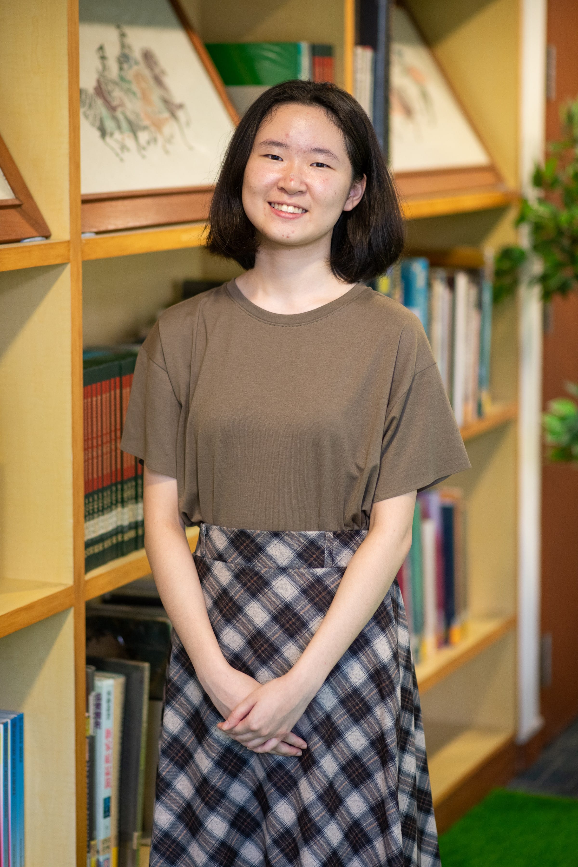 Young woman with short dark hair smiling, wearing a brown shirt and plaid skirt, standing in front of wooden bookshelves.