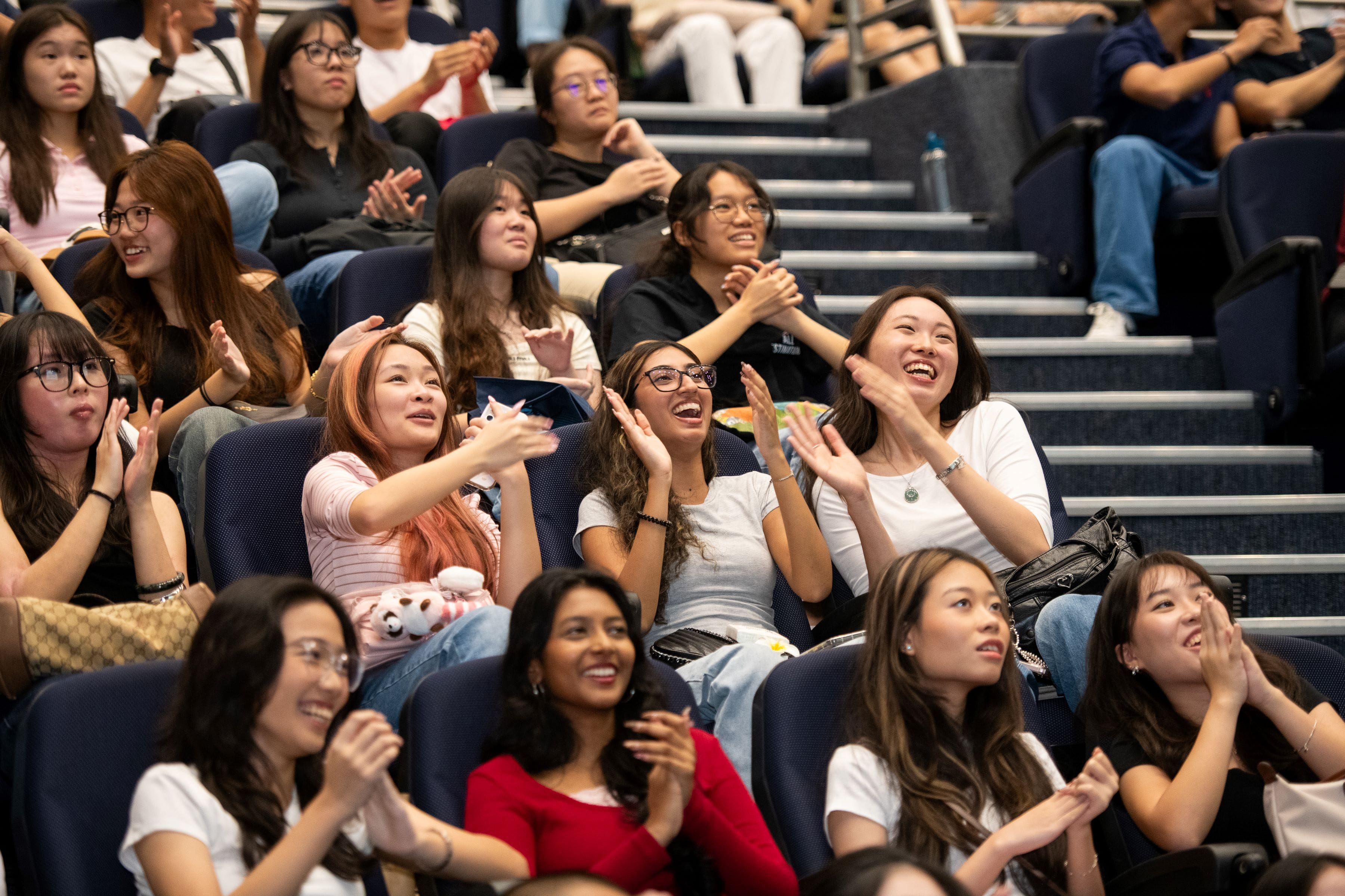 A crowd of young women seated in an auditorium, smiling, clapping, and looking upward with excitement.