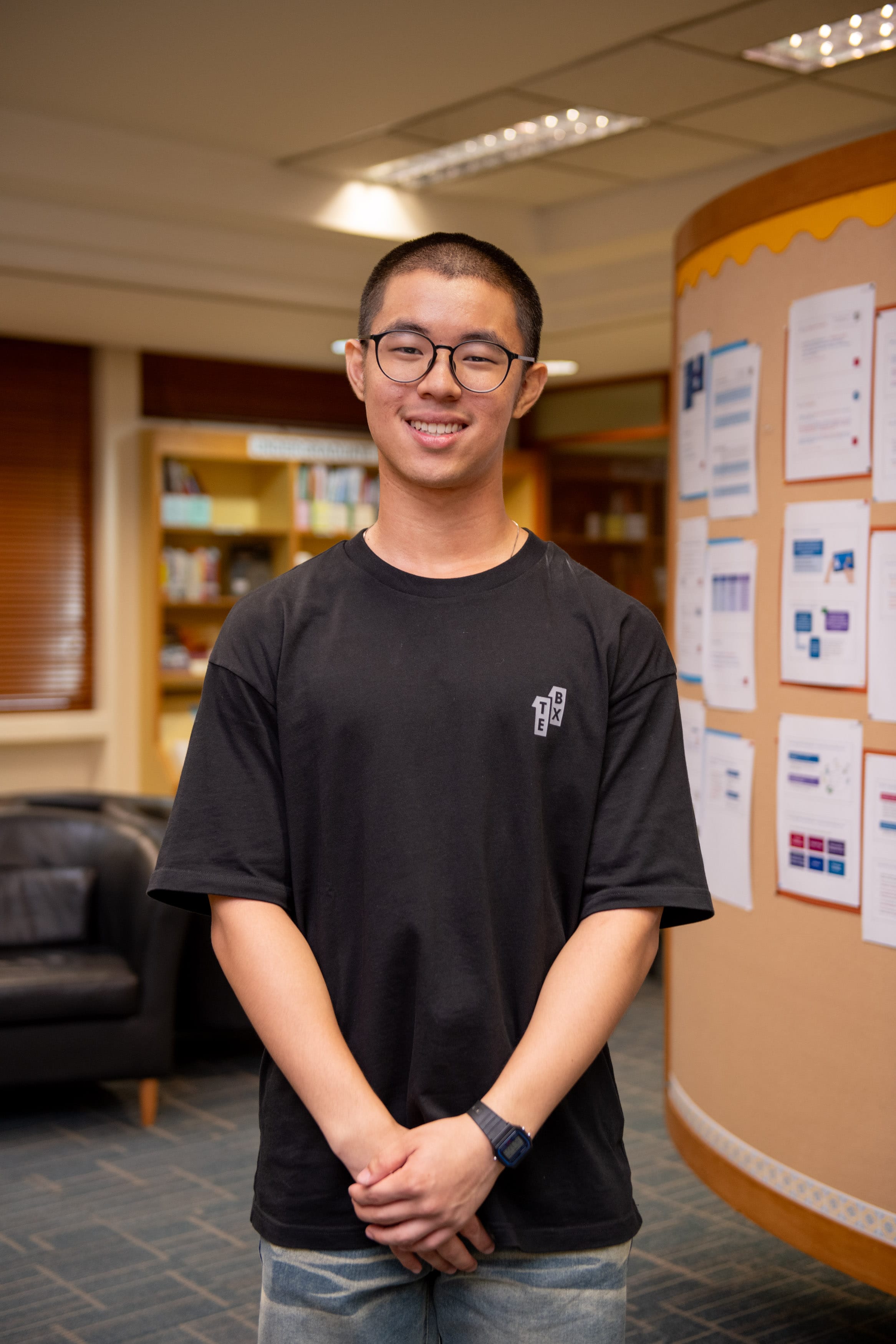 Young man with glasses and short hair, smiling, wearing a black t-shirt, standing in a library near a bulletin board.