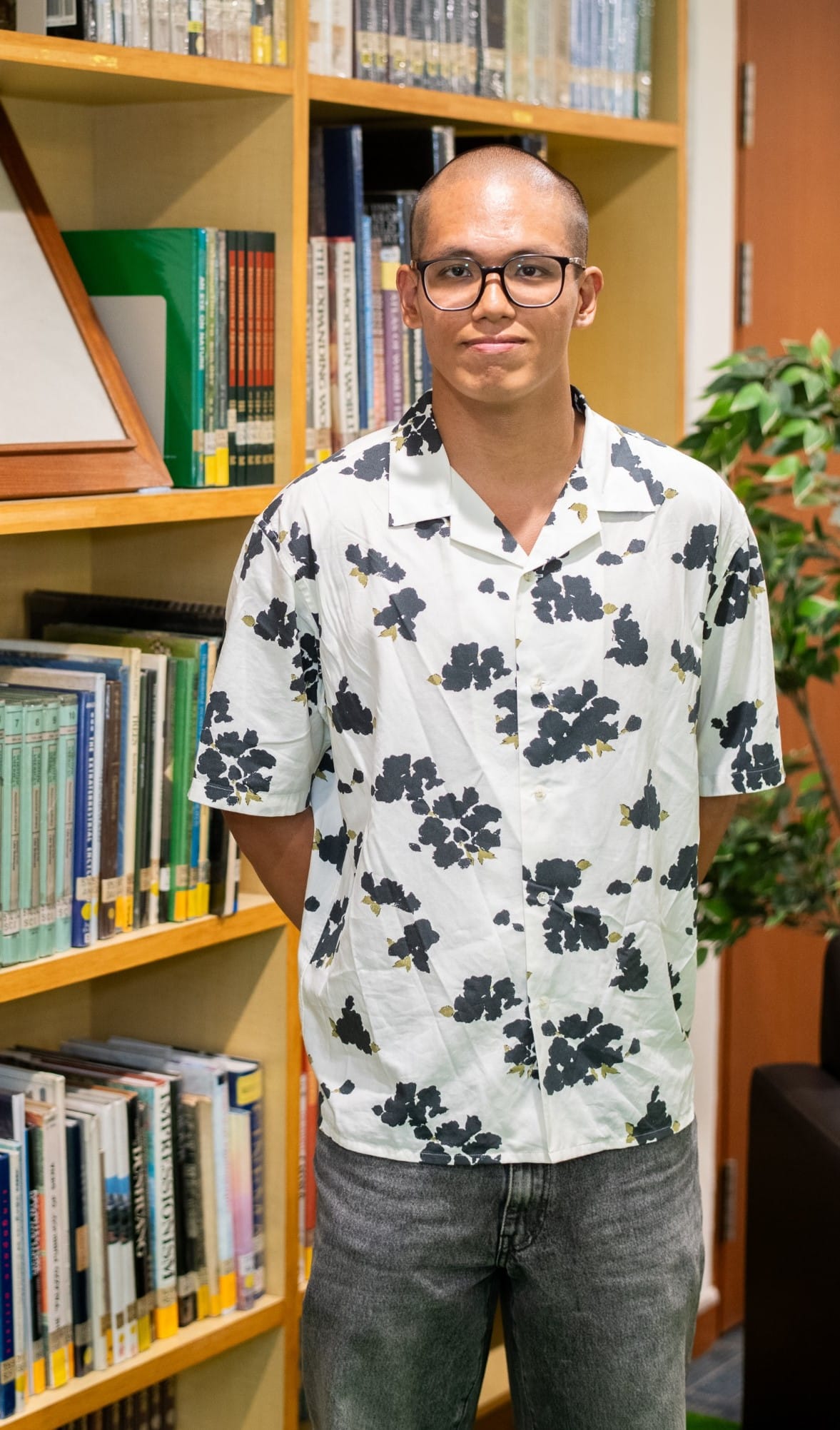 Young man with glasses and a shaved head wearing a white floral shirt stands in front of wooden bookshelves.