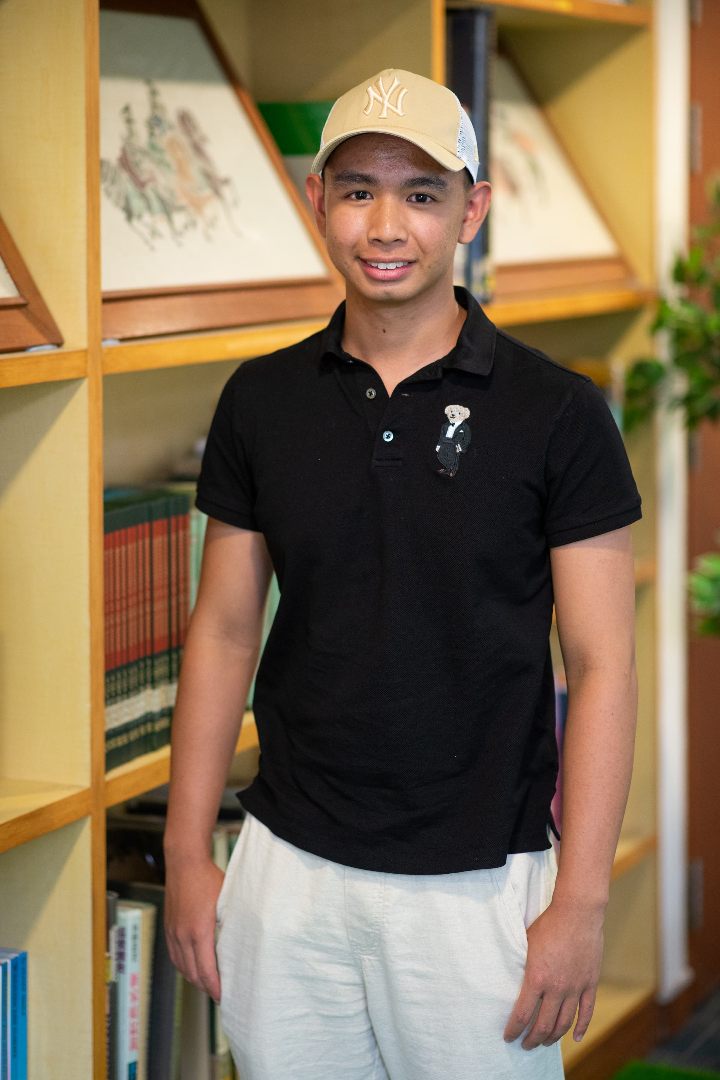 Young man smiling, wearing a black polo shirt and tan NY baseball cap, standing in front of a wooden bookshelf.