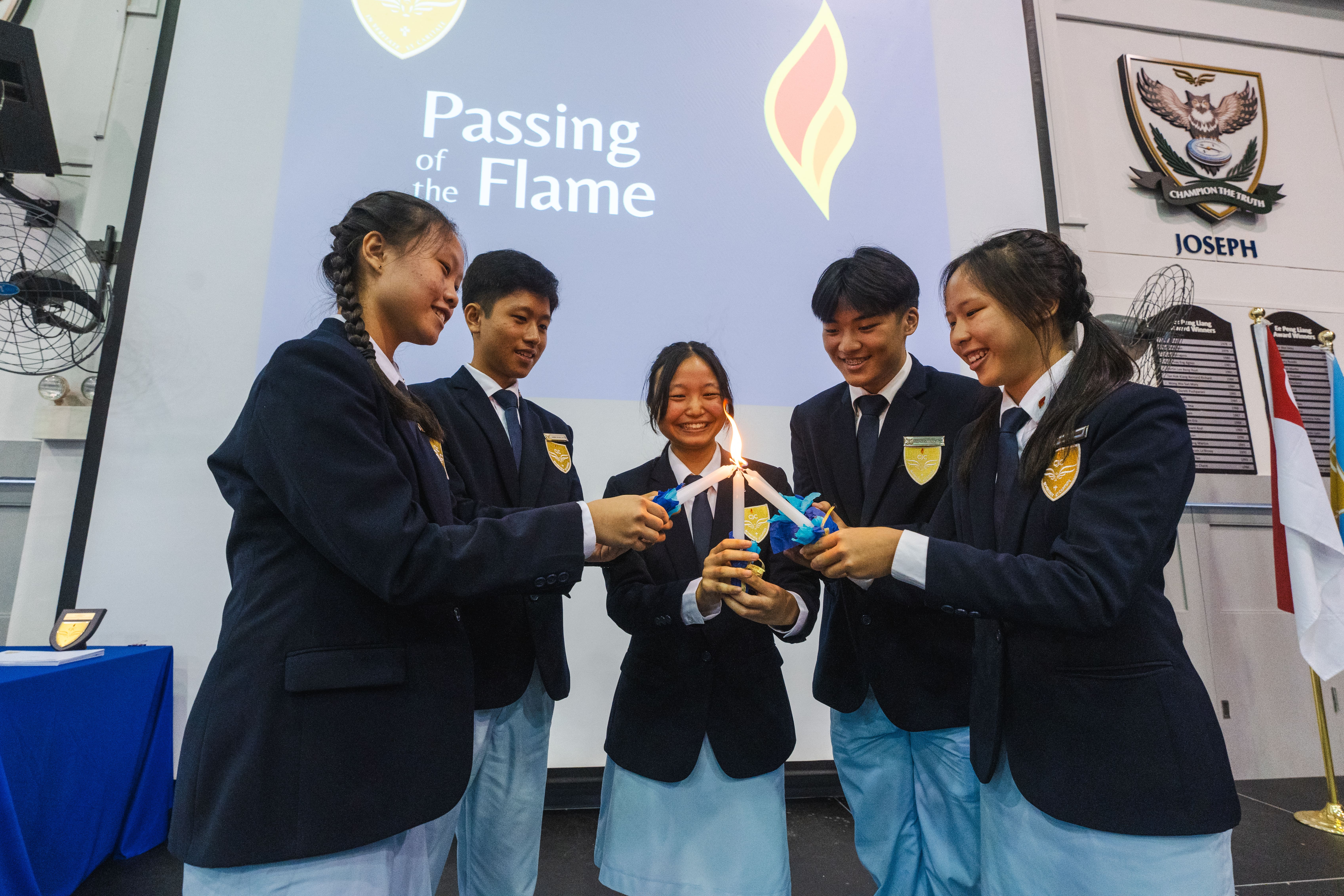 Five students in school uniforms light candles together, smiling under a screen reading Passing of the Flame.