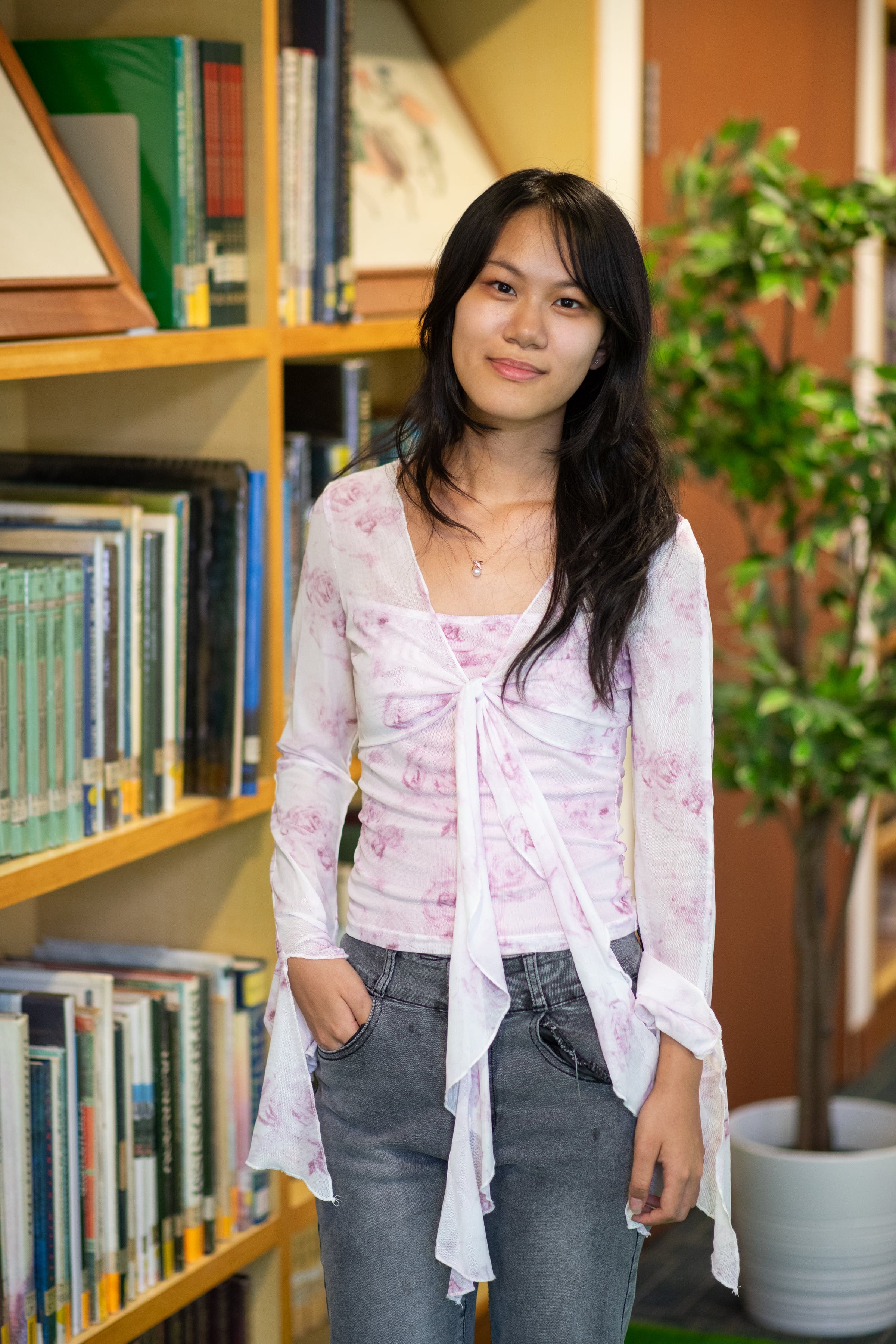 Young woman with long dark hair, wearing a floral top and jeans, smiling in front of library bookshelves.
