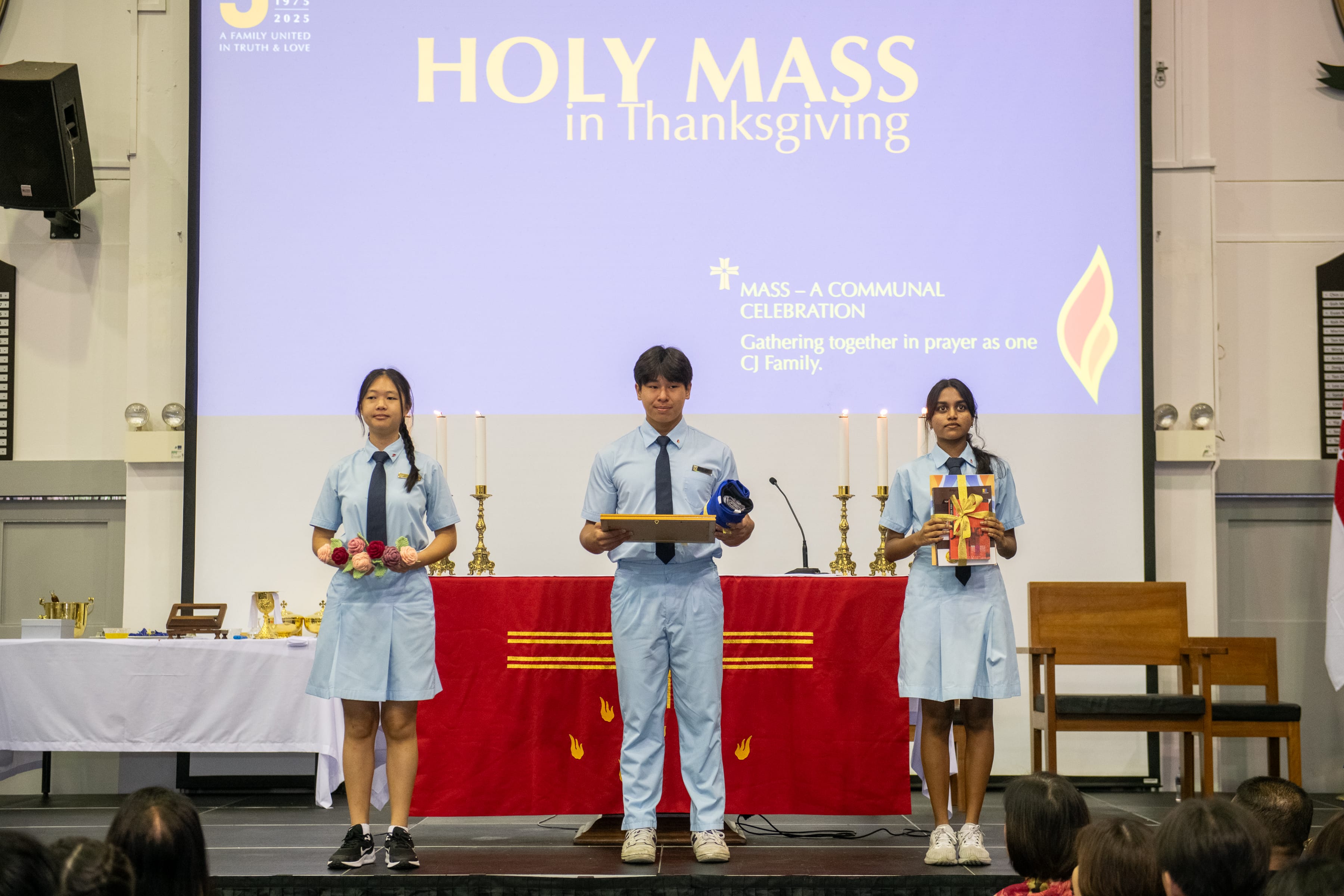 Three students in blue uniforms stand before an altar under a screen reading Holy Mass in Thanksgiving.