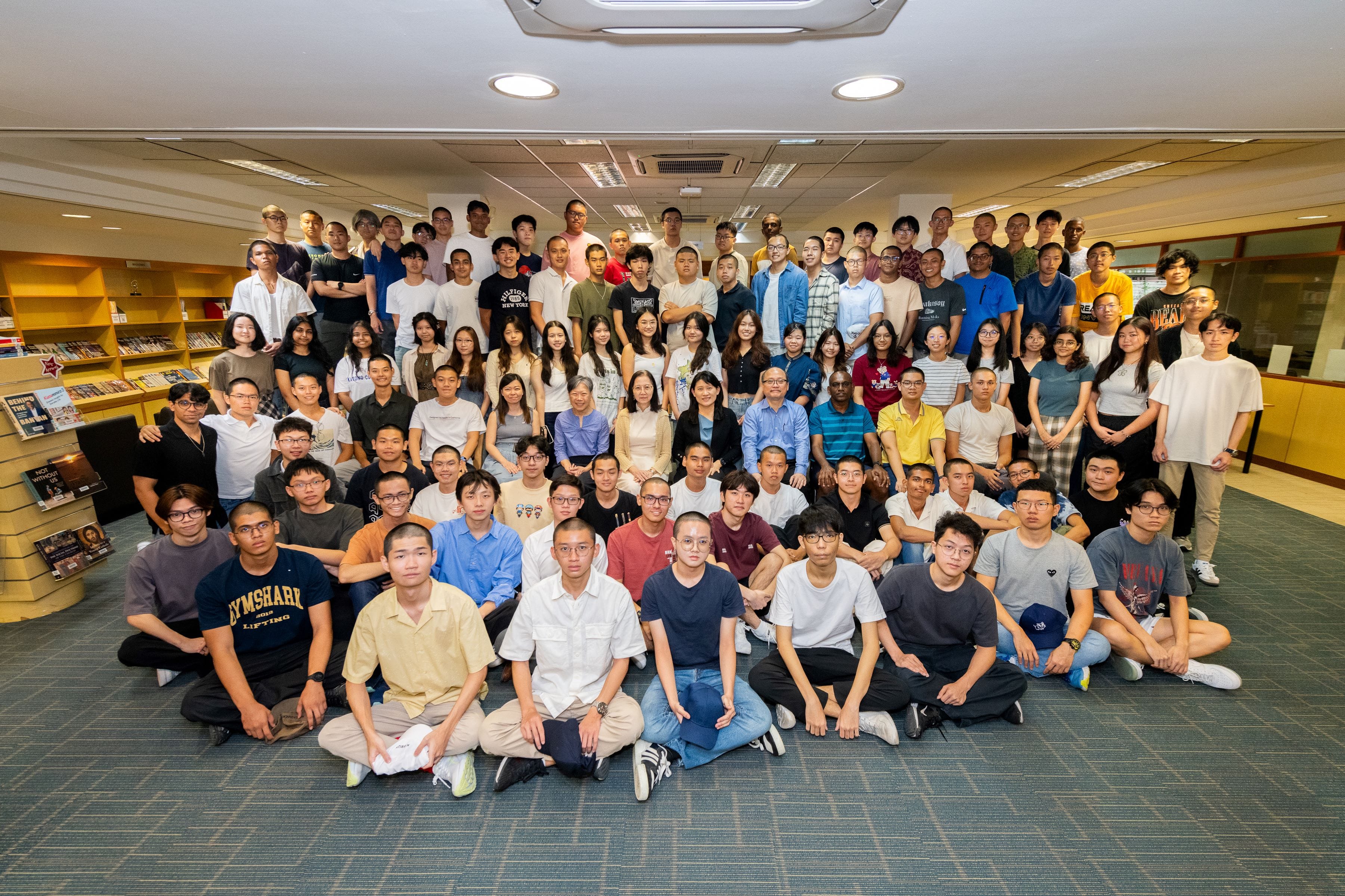 Large group of about 100 people posing together indoors in multiple rows, some sitting on the floor.