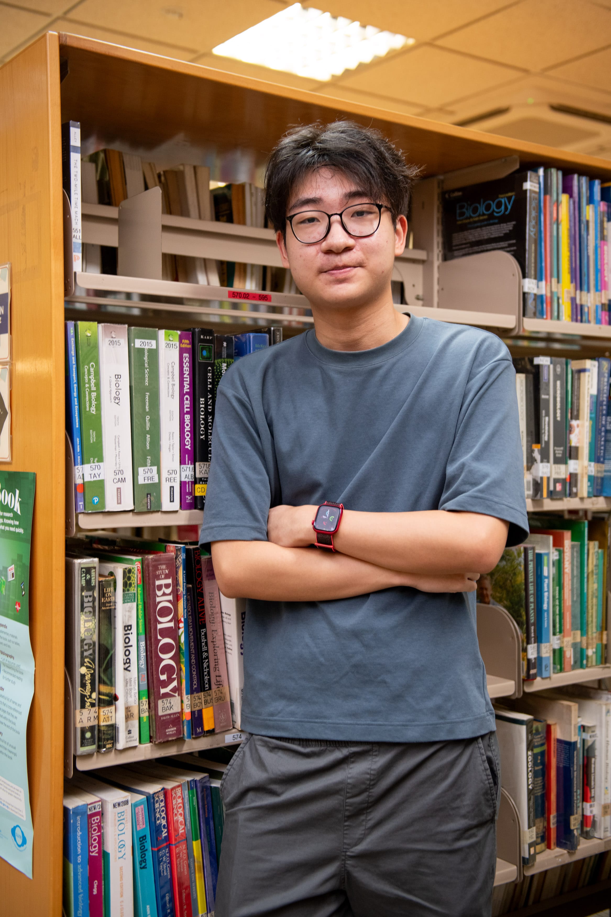 Young man with glasses and a red smartwatch stands arms crossed beside library shelves filled with biology books.