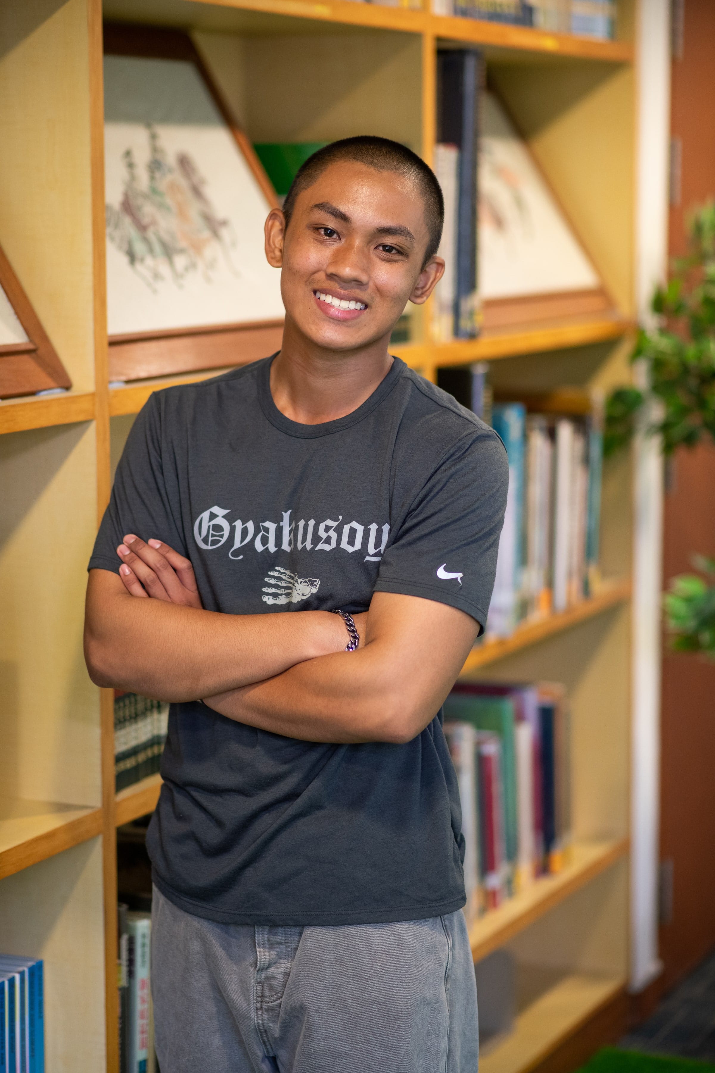 Young man with short hair, smiling with arms crossed, wearing a gray t-shirt, standing in front of wooden bookshelves.