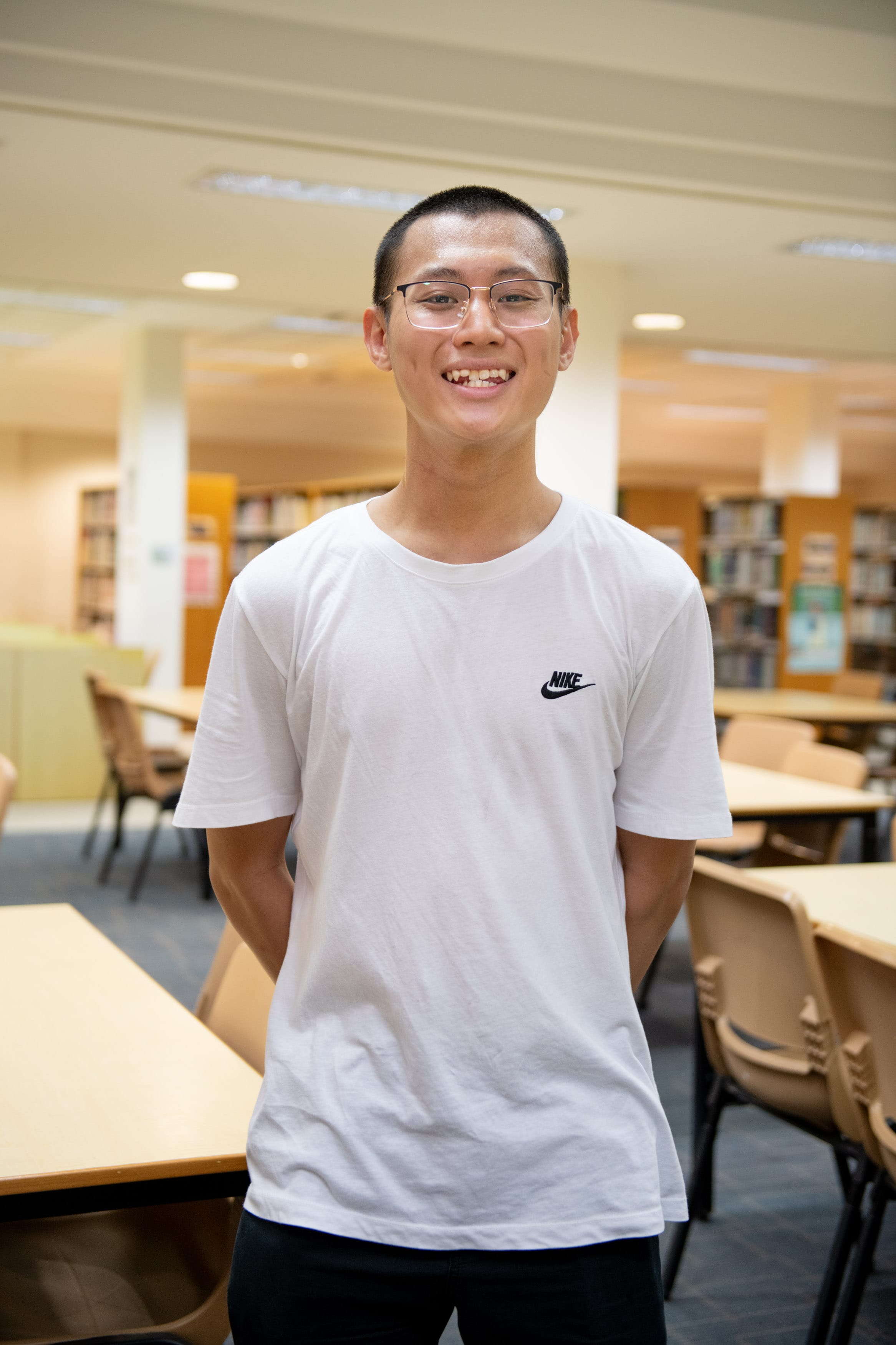 Young man with glasses and short hair smiling, wearing a white Nike t-shirt, standing in a library.