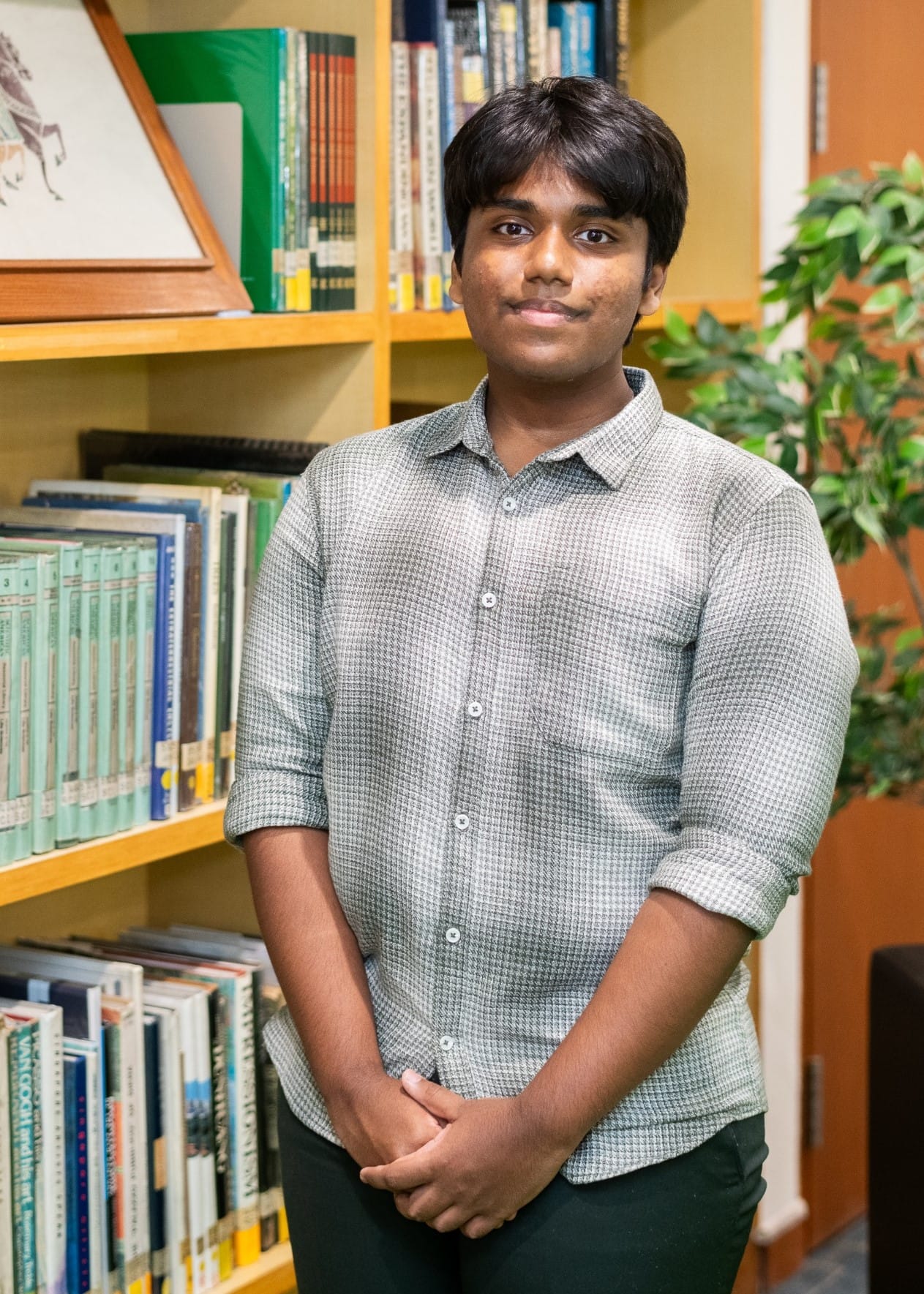 Young man in a gray button-up shirt stands smiling in front of wooden bookshelves filled with books.