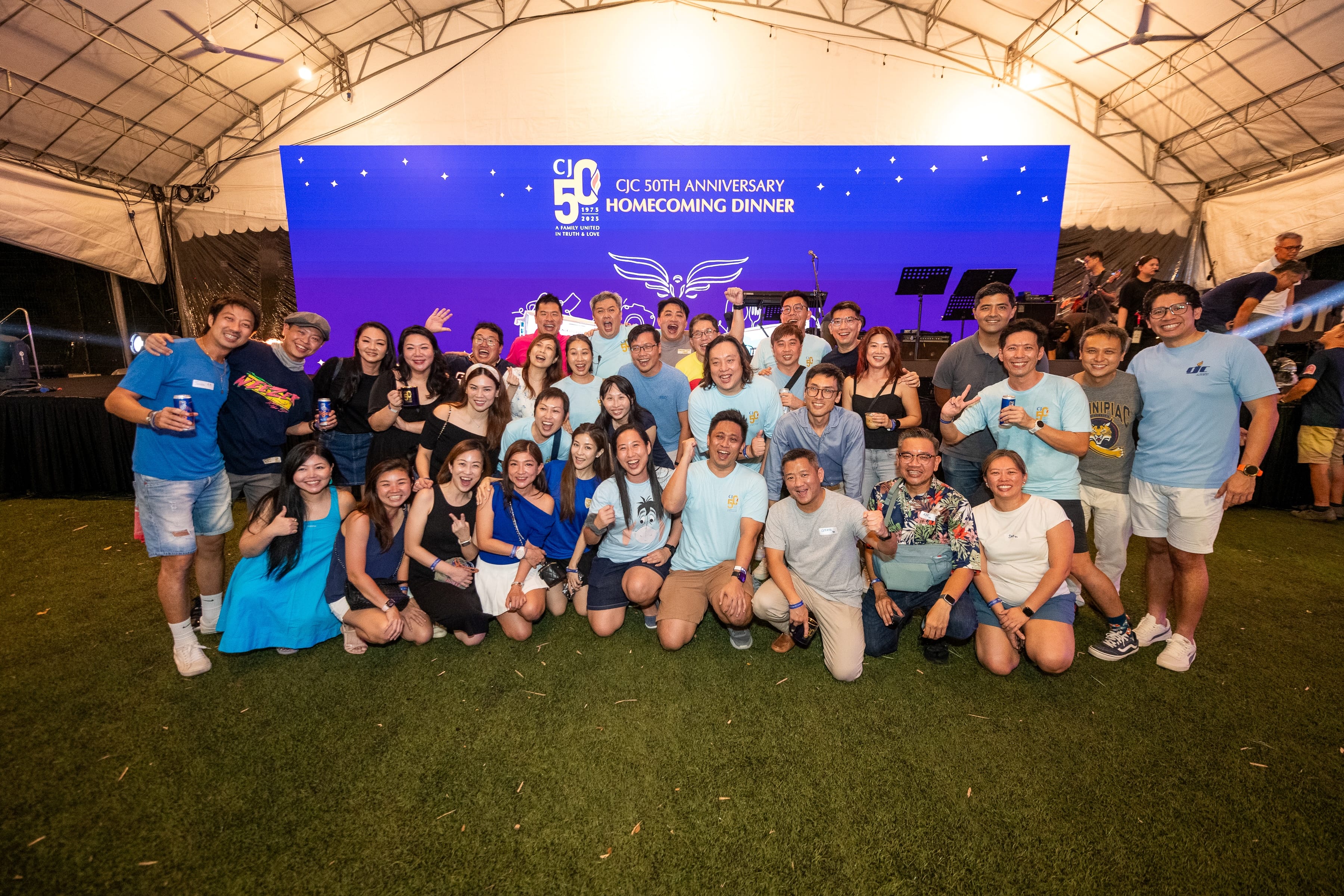 Large group posing on grass before stage backdrop reading CJC 50th Anniversary Homecoming Dinner