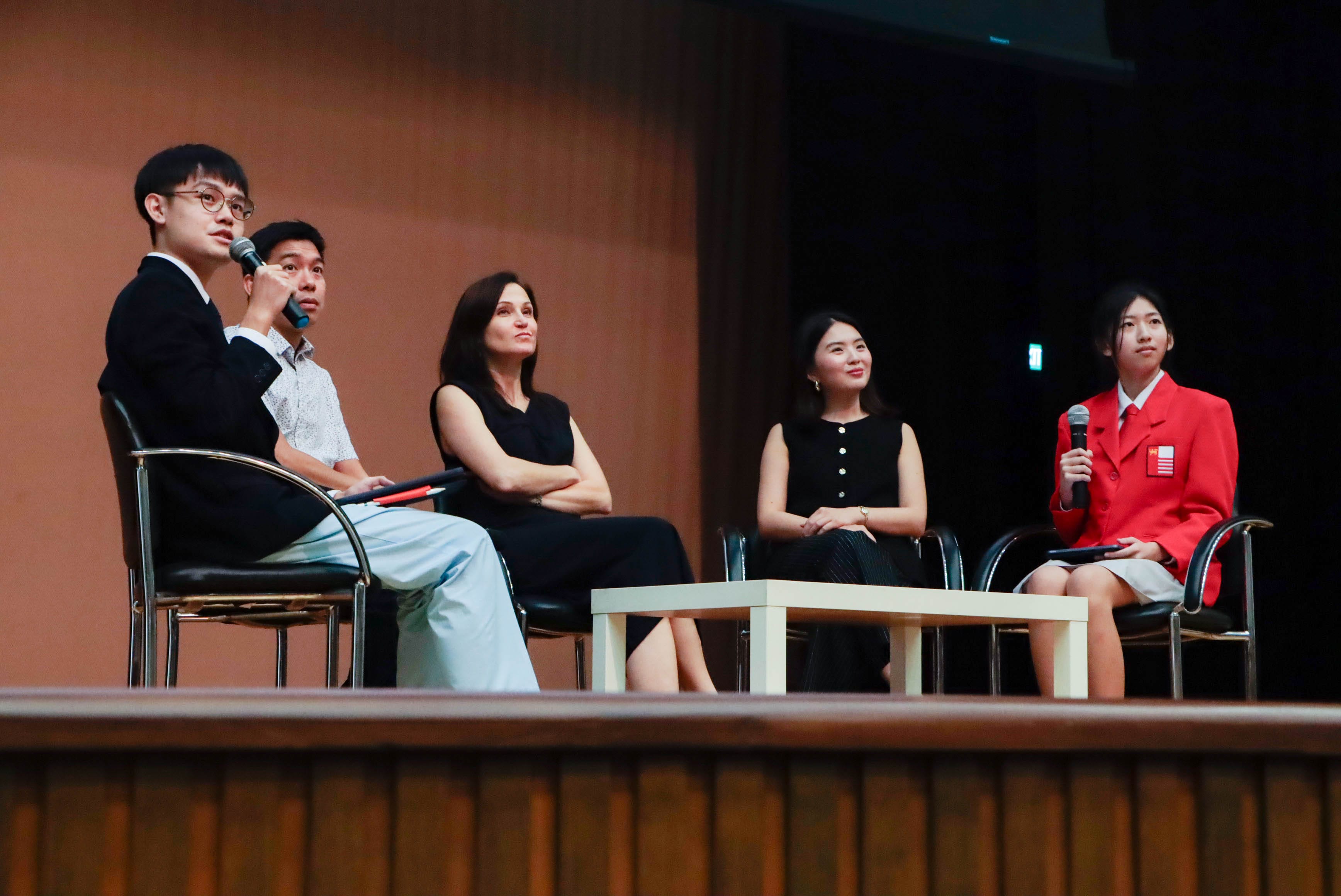 Five people sit on chairs on a stage; a man in glasses speaks into a microphone while others listen.