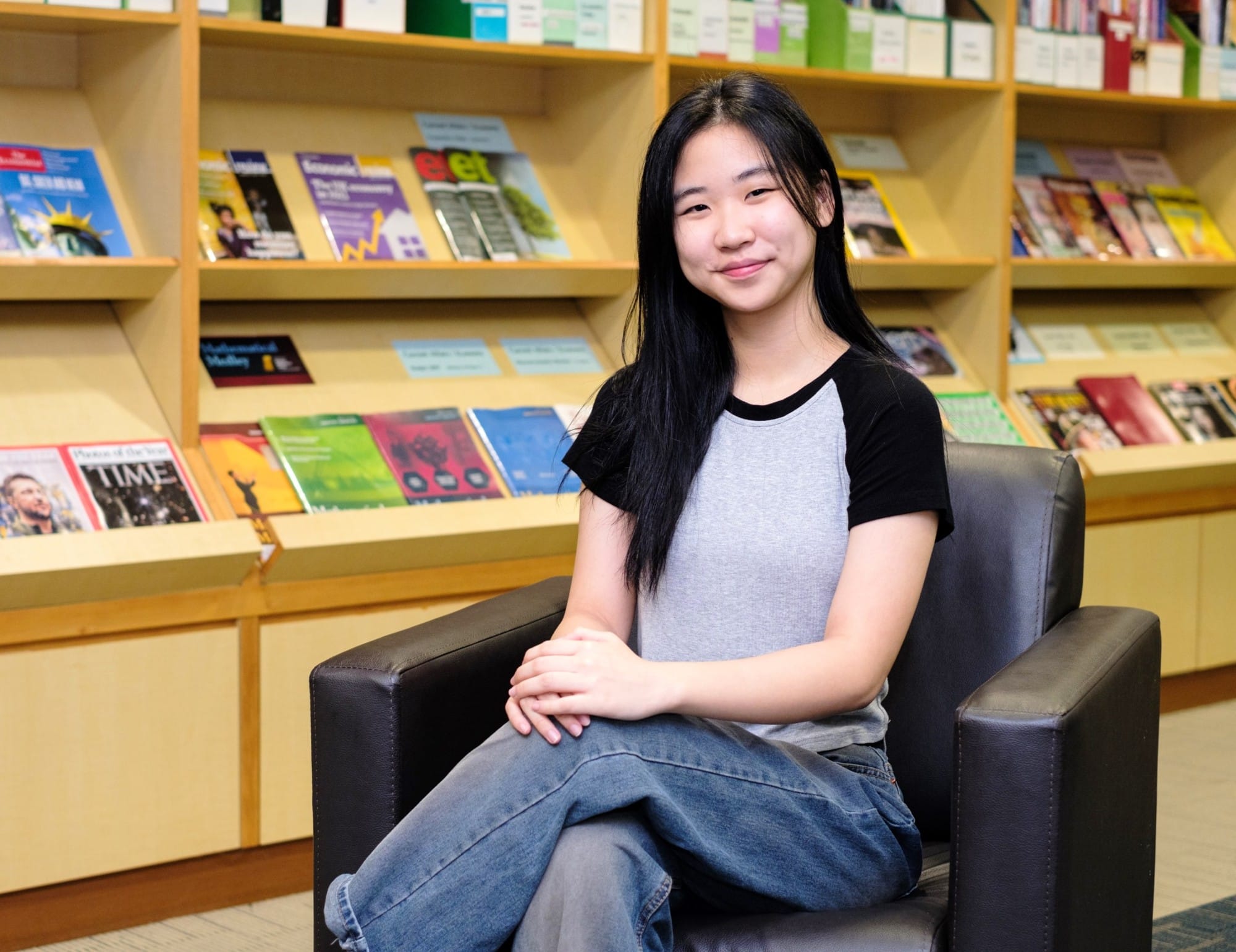 Young woman with long black hair sits smiling in a dark armchair before shelves of colorful magazines.