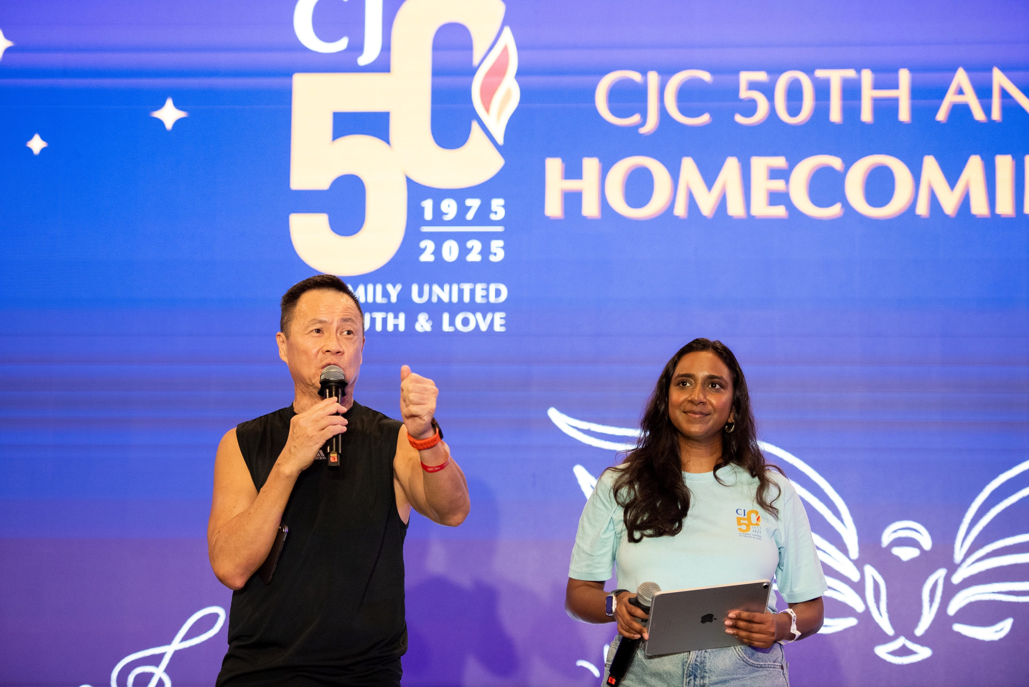 Man speaks into a microphone beside a woman holding a tablet, in front of a CJC 50th anniversary backdrop.