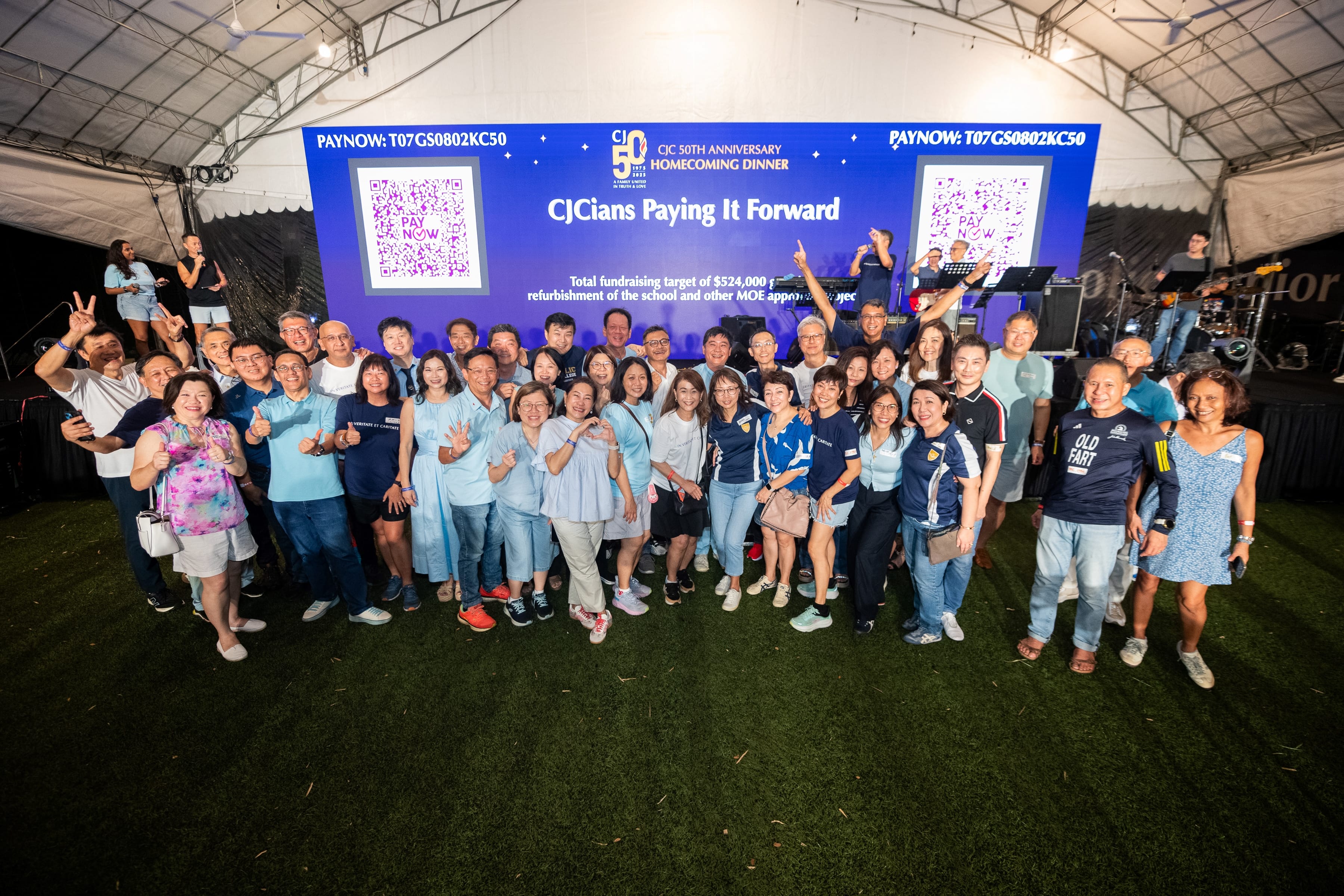 Large group posing under a tent in front of a screen reading CJCIans Paying It Forward, with QR codes.