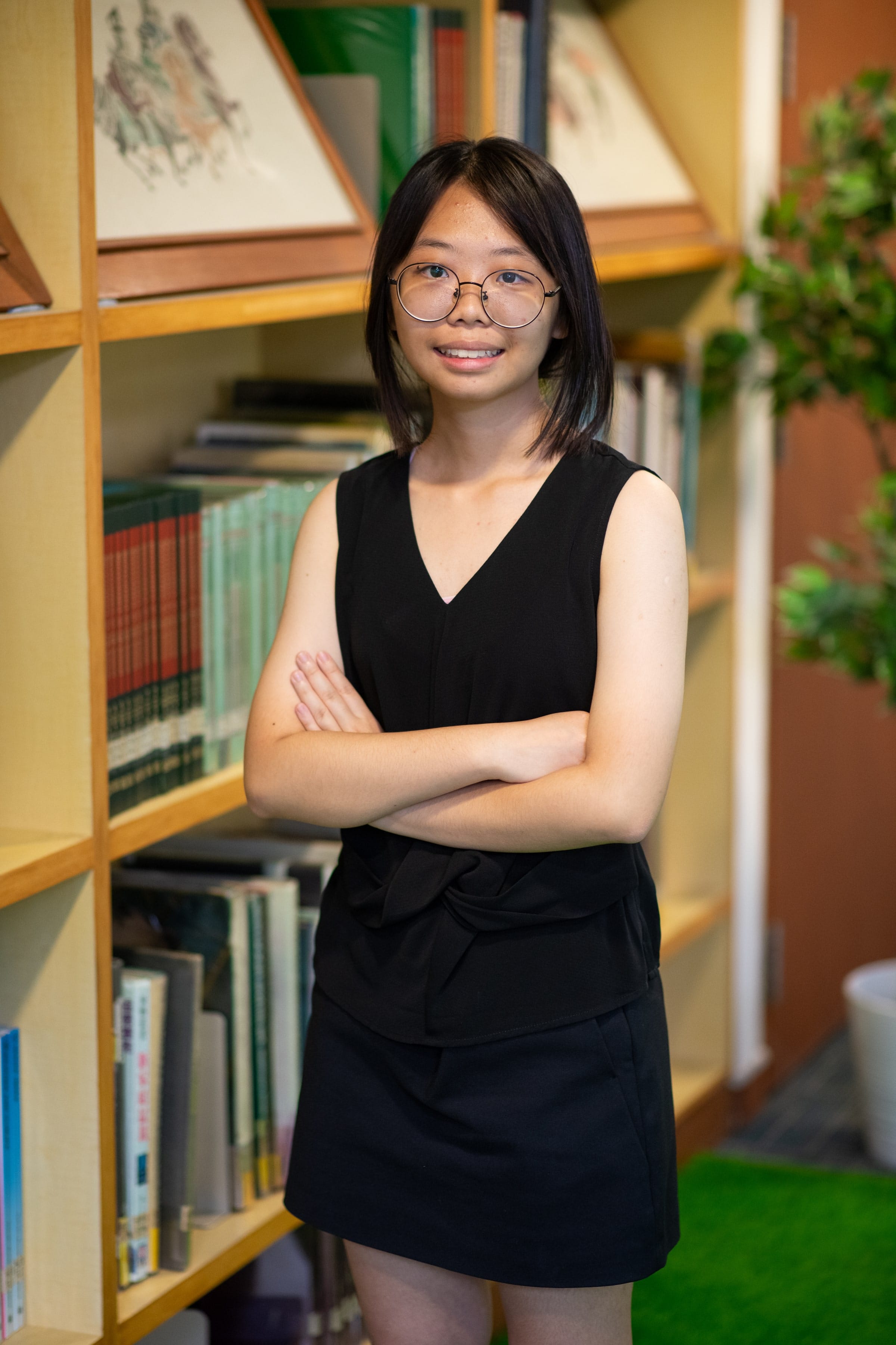 Young woman with glasses and black dress stands smiling with arms crossed in front of wooden bookshelves.