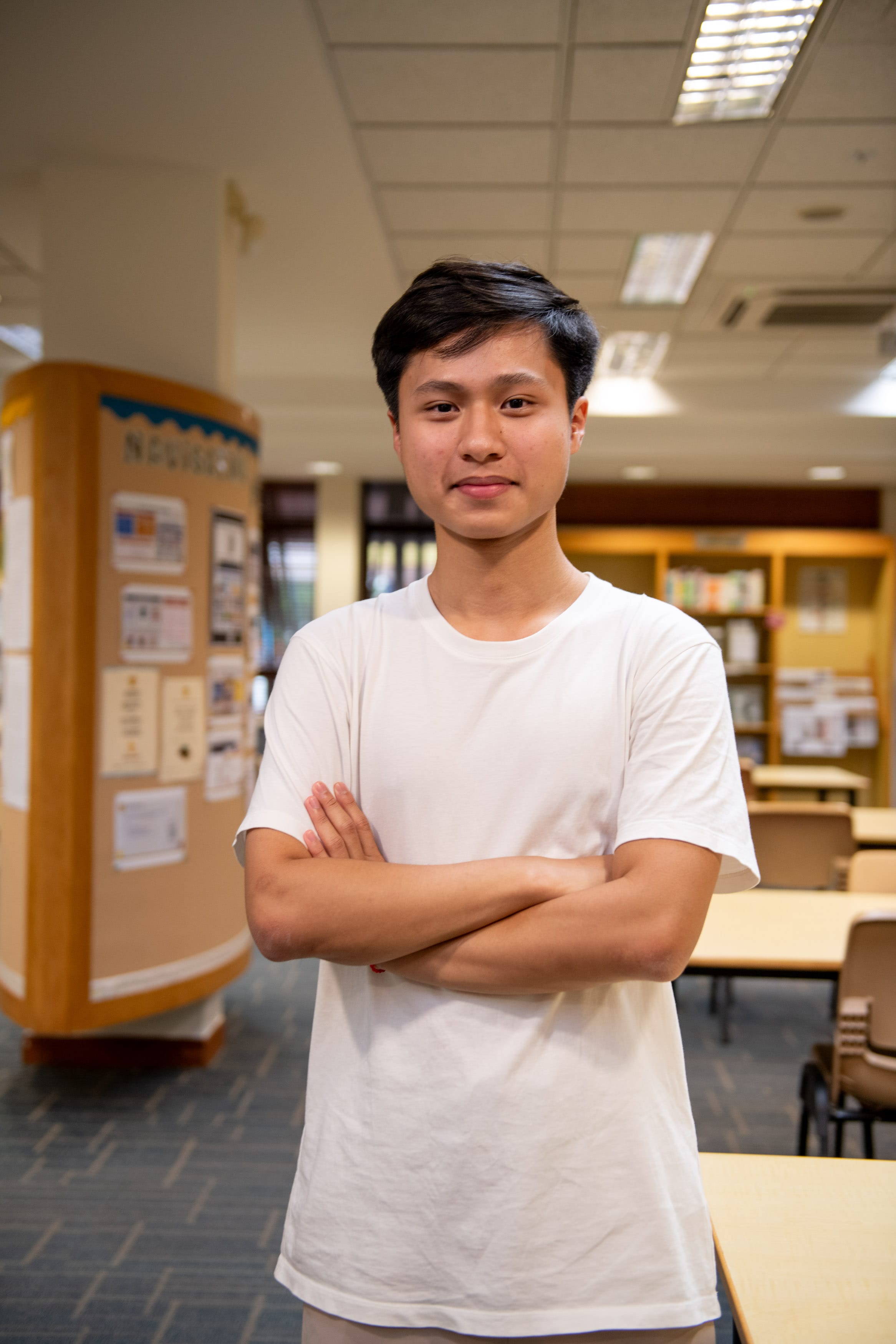 Young man in a white t-shirt standing with arms crossed, smiling in a library with bookshelves and tables behind him.